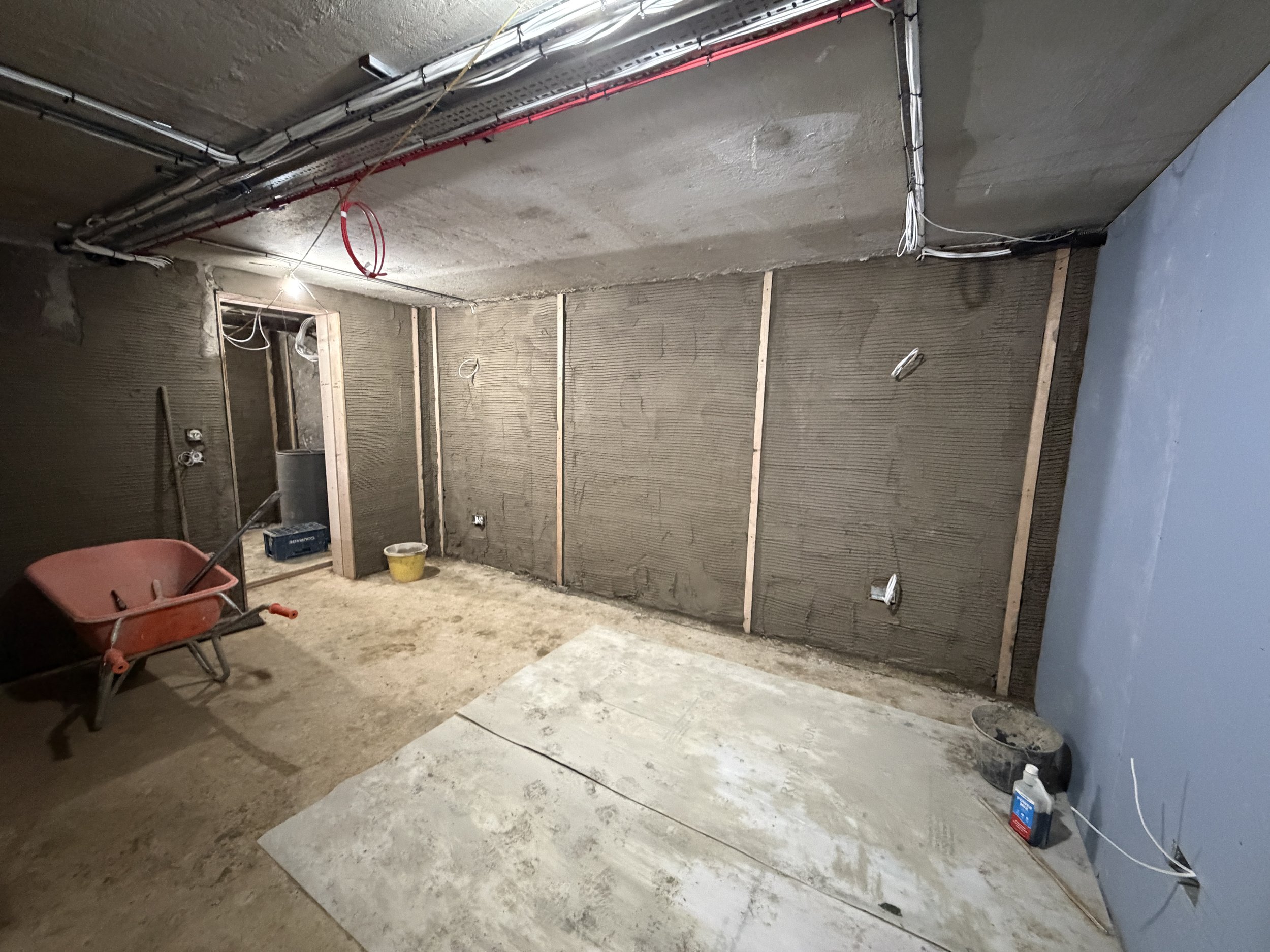 Interior of a room under construction with exposed wiring, framing, and unfinished walls. There is a wheelbarrow, a bucket, and construction materials on the floor.