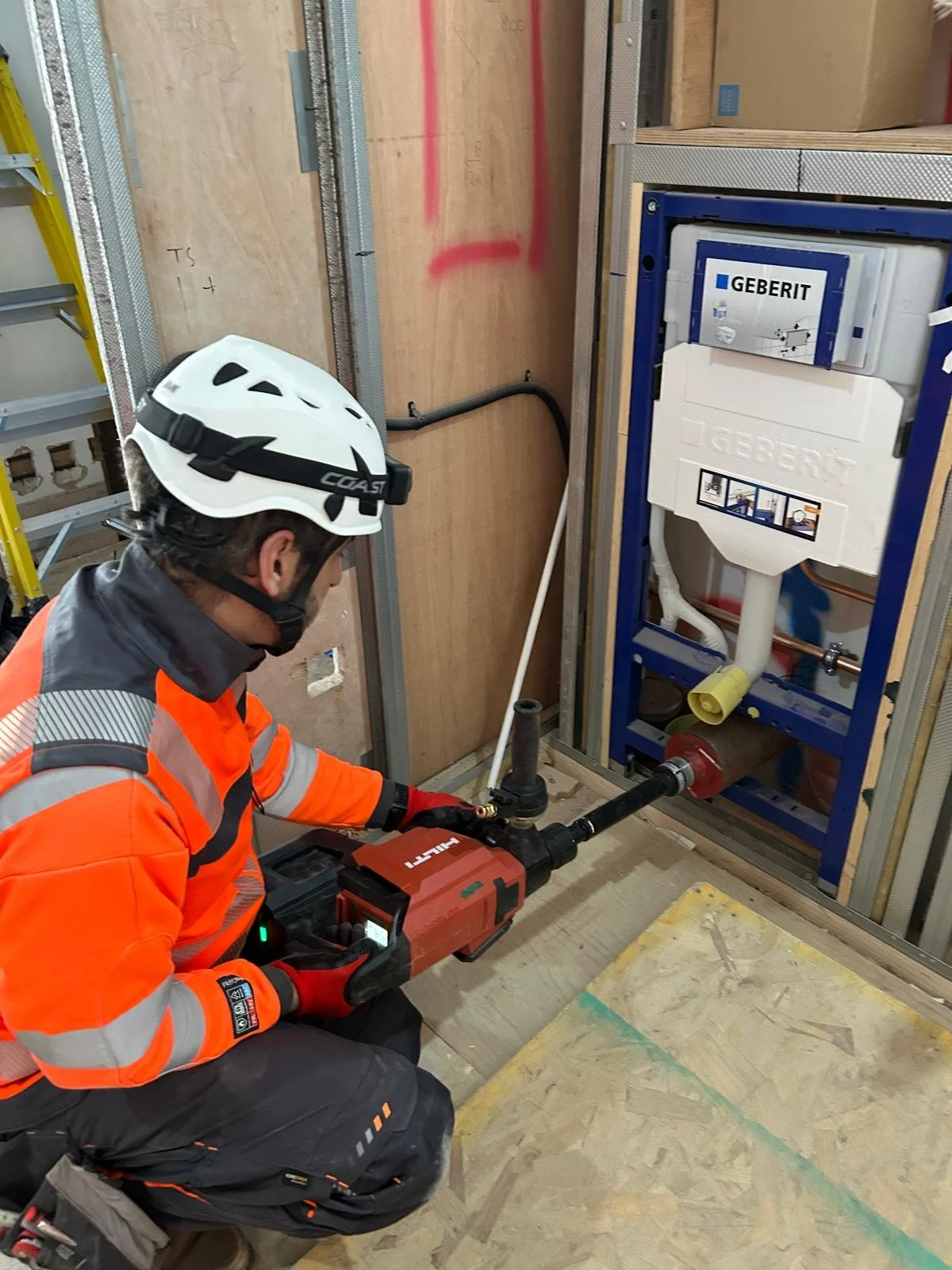 A construction worker wearing an orange safety jacket, helmet, and gloves kneels while inspecting plumbing work in a partially built wall.