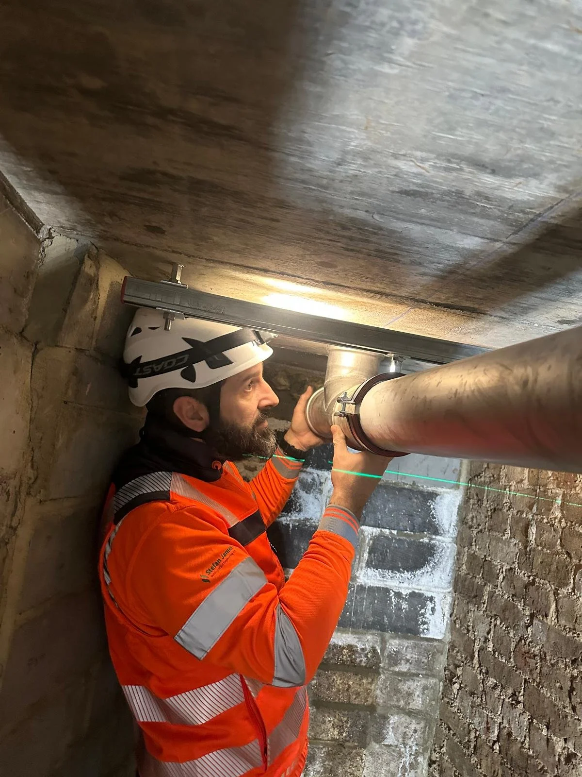 A worker in an orange safety jacket and white helmet inspects a pipeline in a brick-walled underground tunnel using a laser measuring device.