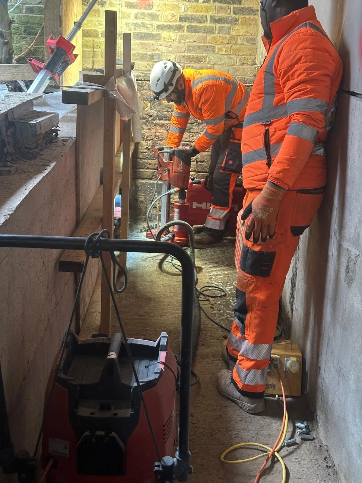 Two construction workers in orange safety suits and helmets working inside a narrow room with brick walls. One worker is handling a power tool, while the other stands nearby. Various tools and equipment are visible around them.