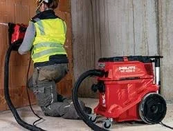 A person wearing a yellow safety vest kneels on the floor using a wet vacuum cleaner to clean a surface.
