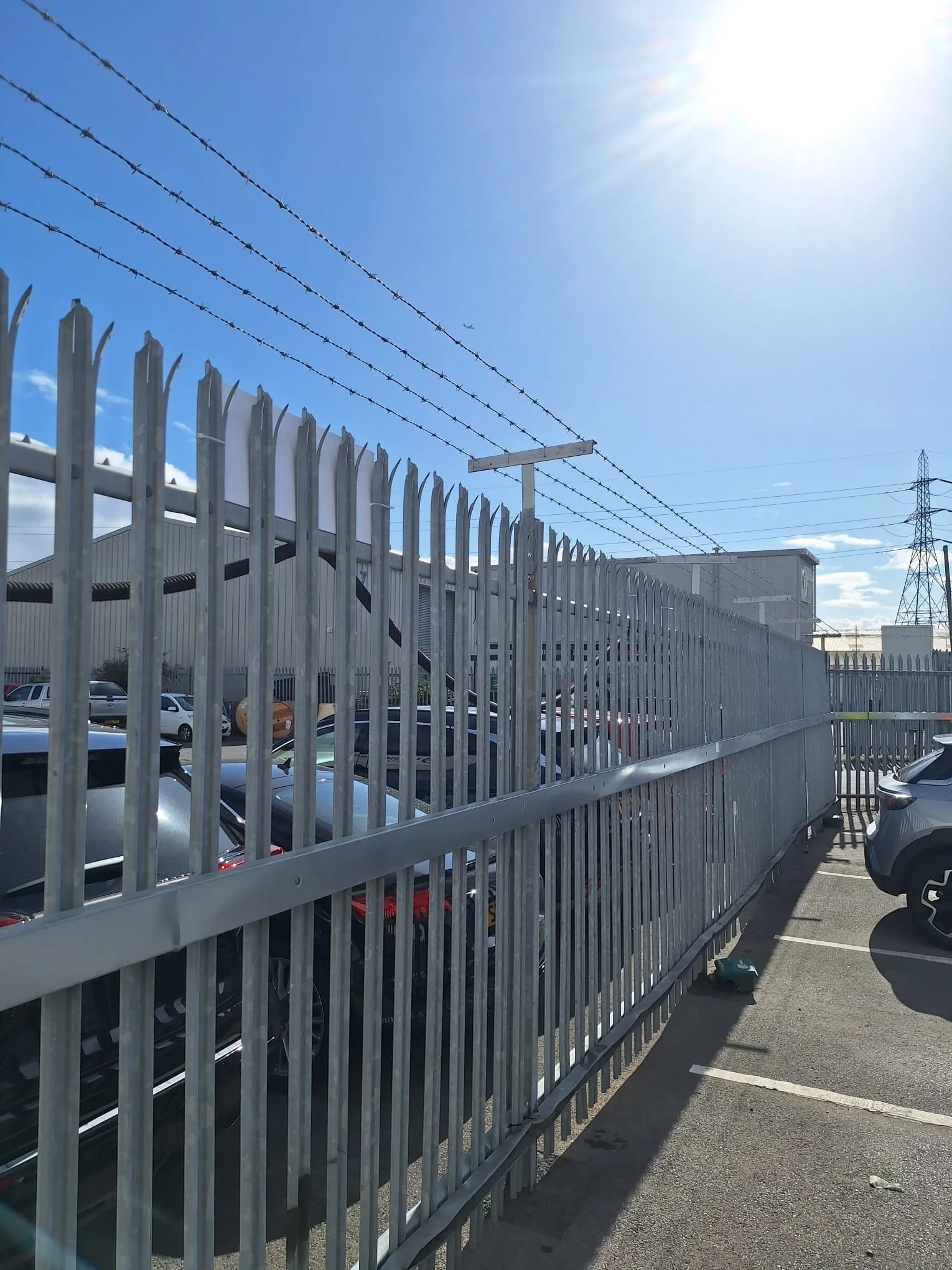 A tall metal security fence topped with barbed wire runs along a parking lot, with several parked cars underneath and a building in the background. A large airplane is visible flying in the blue sky. The sunlight is bright, creating glare in the top right corner of the image.