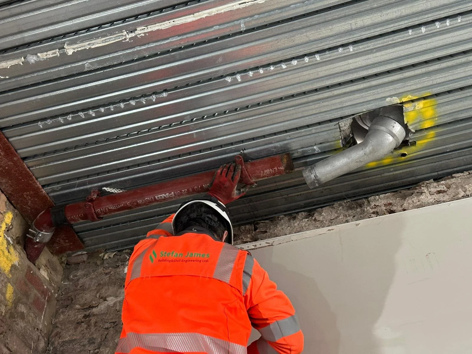 A construction worker in an orange vest and helmet is working on a wall, installing or inspecting plumbing pipes near a metal ceiling with exposed pipes and ductwork.