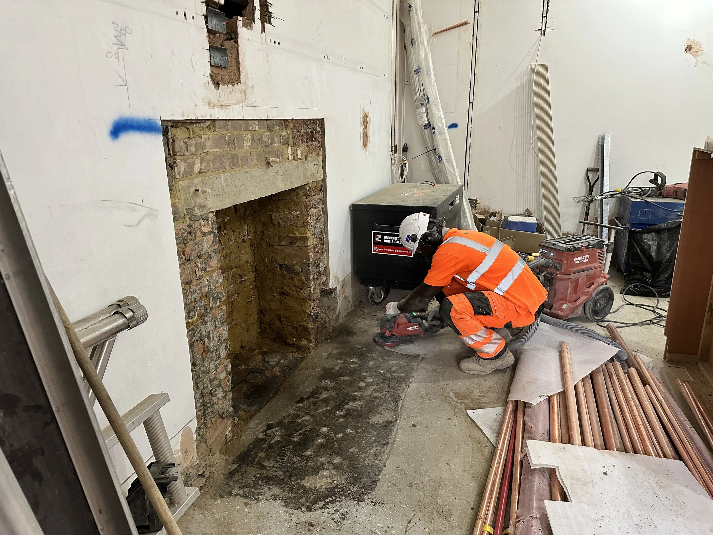 A construction worker wearing an orange safety vest and helmet working on a renovation project. The room has exposed brick and drywall with some pipes visible, construction tools, and equipment scattered around.