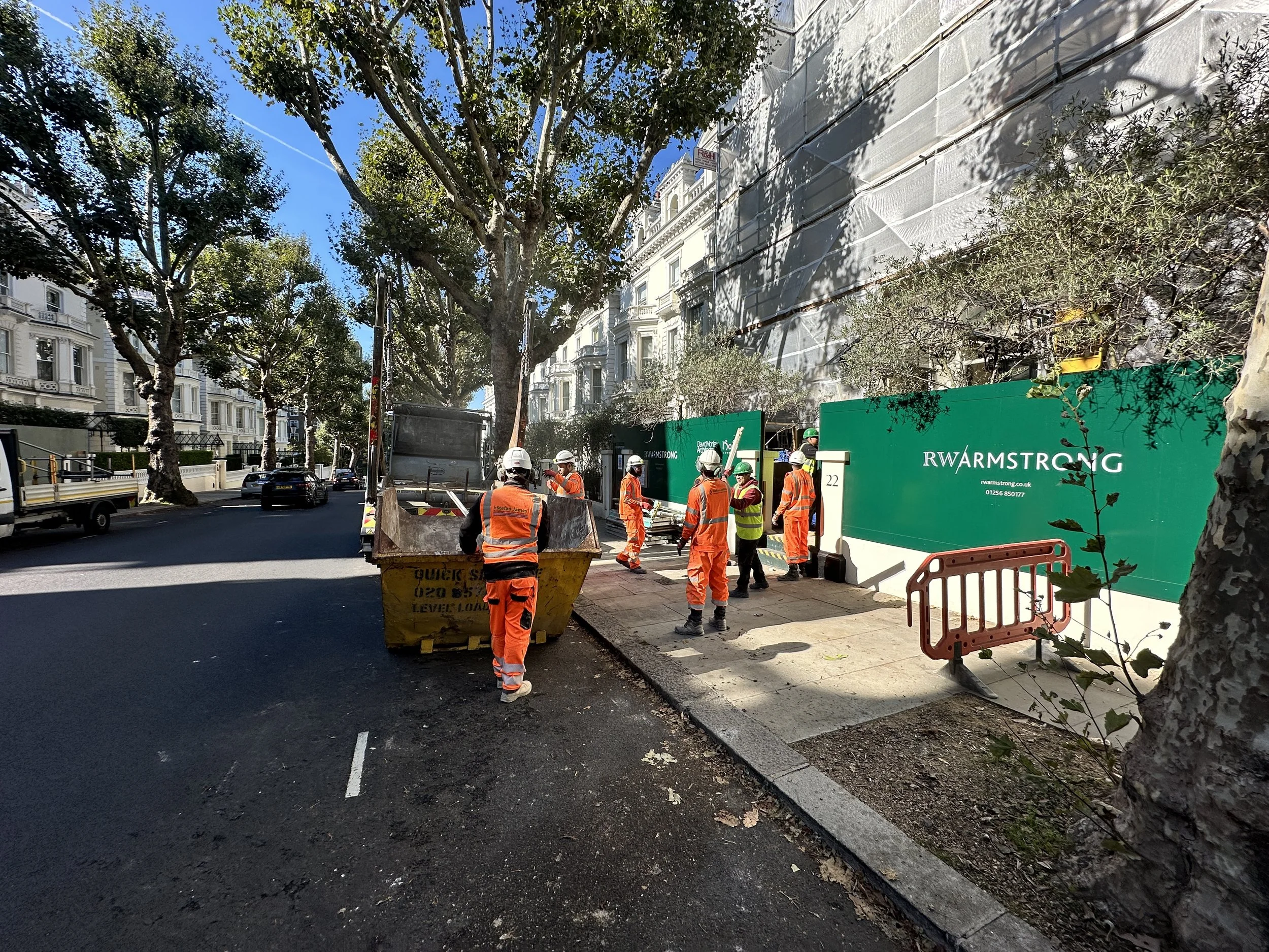 A street construction scene with workers in orange safety vests and white helmets working on the sidewalk. There are trees lining the street and large white buildings in the background. Green barriers with text are set up around the work area.