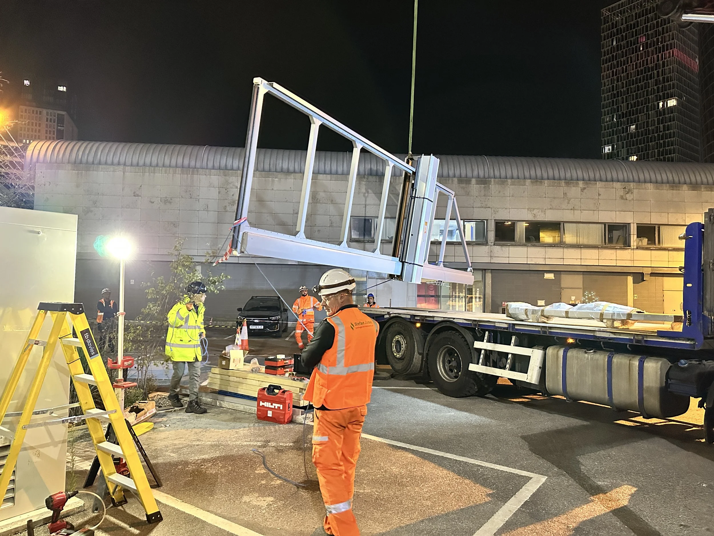 Construction workers in safety vests and helmets working at night on a large construction project involving a crane truck and equipment, with a modern building in the background.