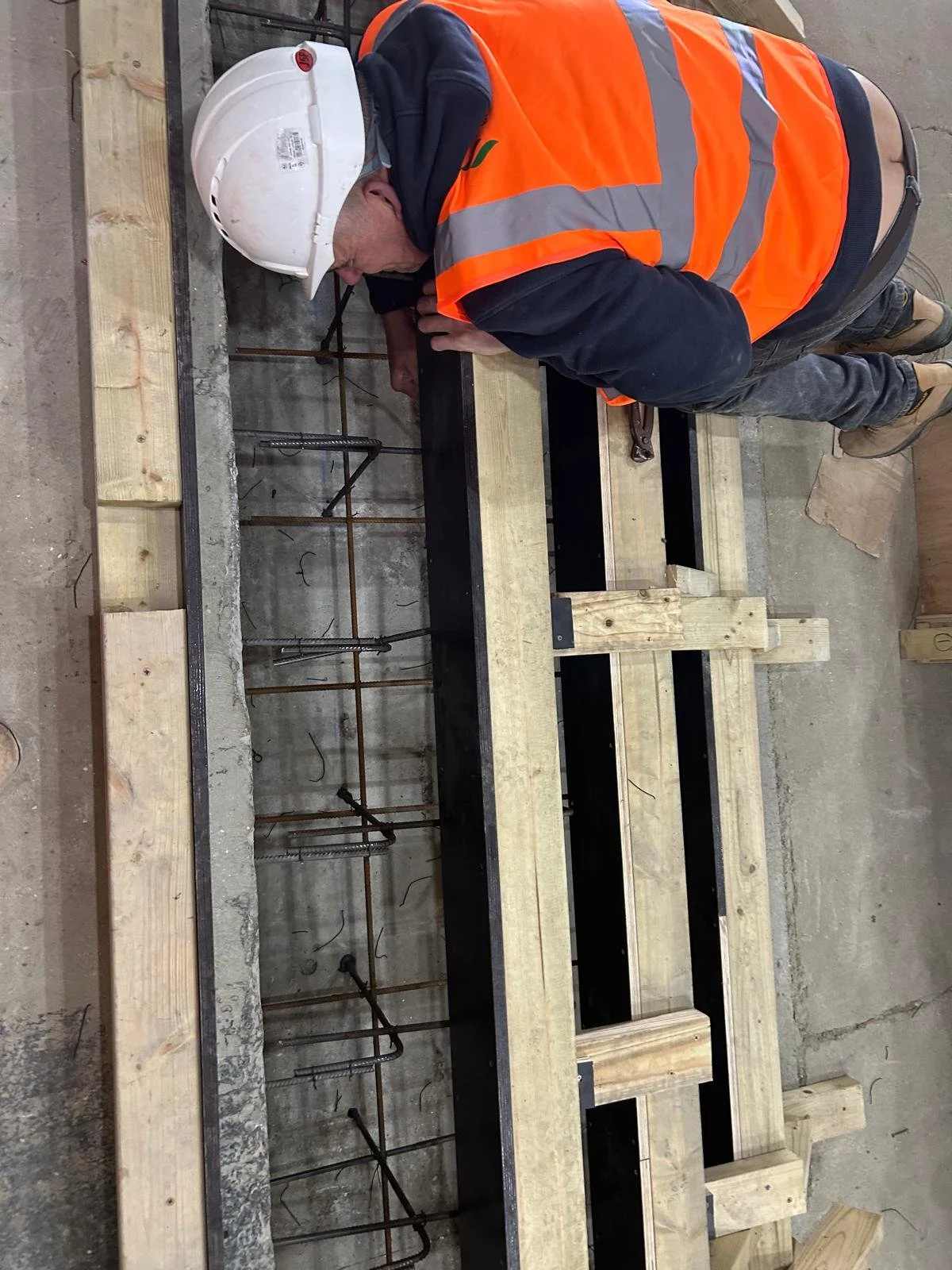 A construction worker wearing a white safety helmet and orange safety vest bent over a work table, working on a wooden frame with exposed metal rebar underneath.