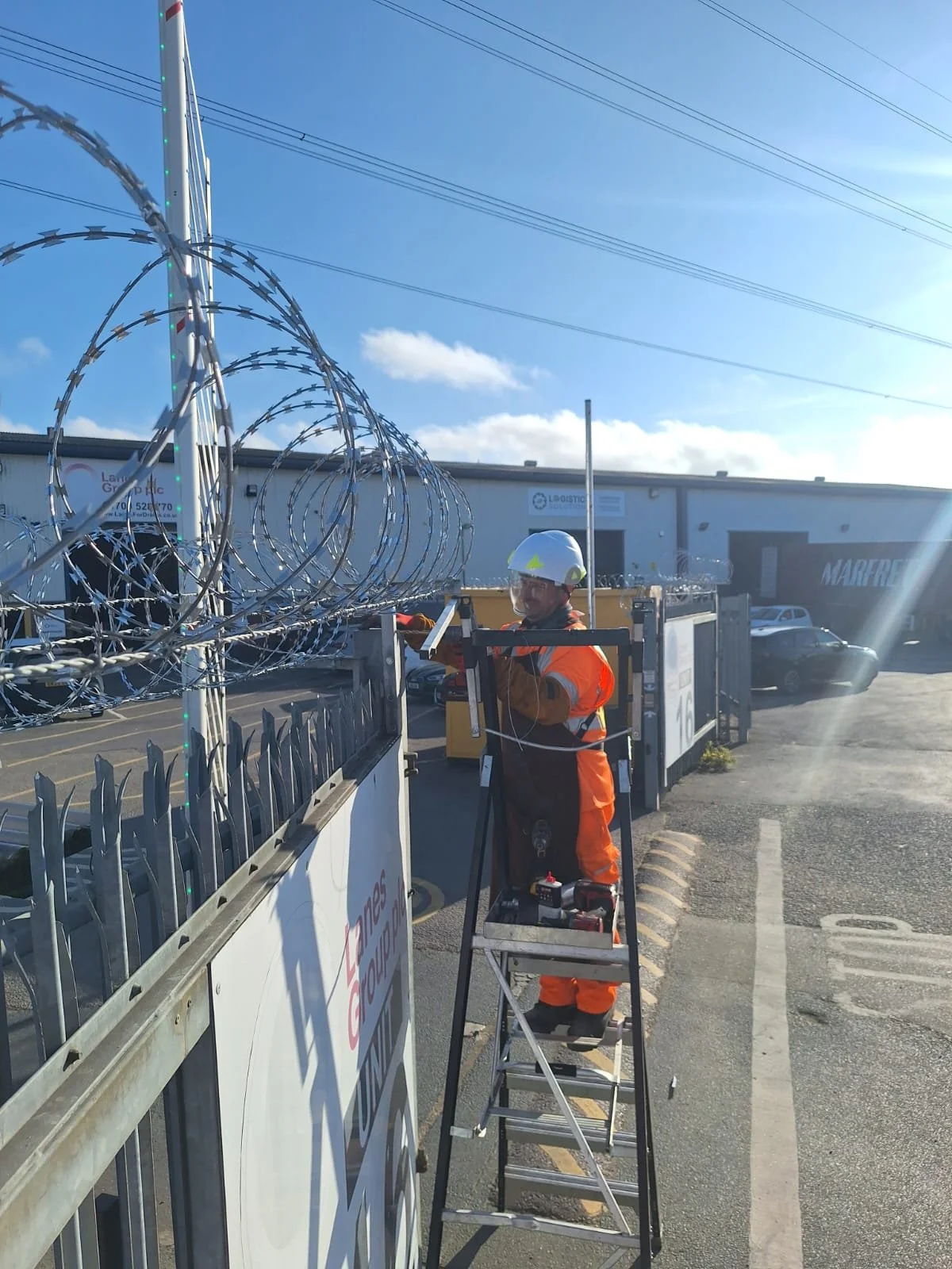 A worker wearing an orange safety jacket and helmet is using a tripod-mounted device to work on fencing topped with spiral coil razor wire outside a commercial building. The sky is mostly clear with some clouds, and there are parked cars and a building in the background.