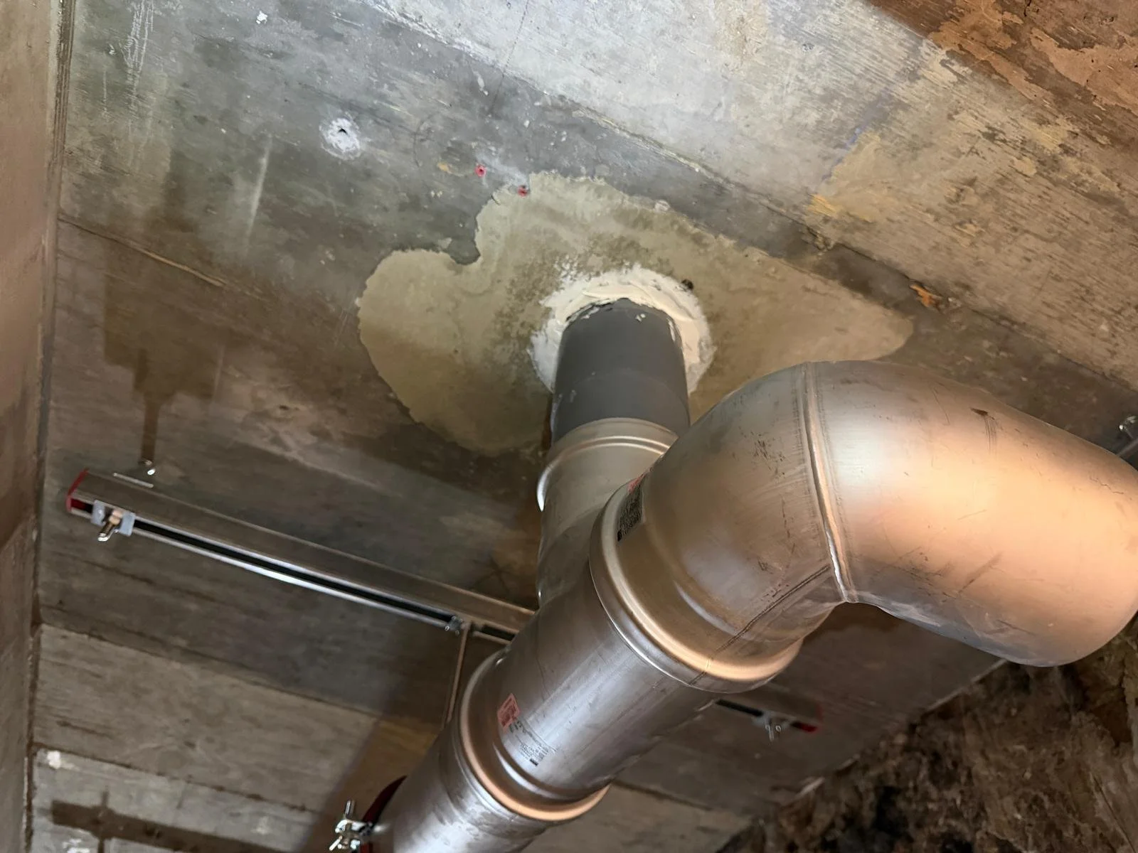 A metal pipe connected to a black fitting is installed through a concrete ceiling, with a circular patch of sealant or mortar around it. The ceiling around the pipe is unfinished, with visible wood framing and subflooring.