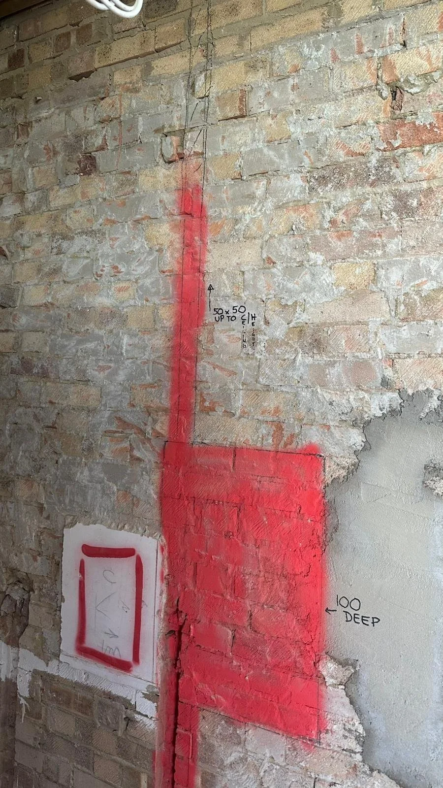 A brick wall with red spray-painted markings, including a vertical line with a 50x50 inch measurement and an arrow pointing upward, a large red rectangle indicating a 100-inch deep section, and handwritten notes with measurements and annotations.