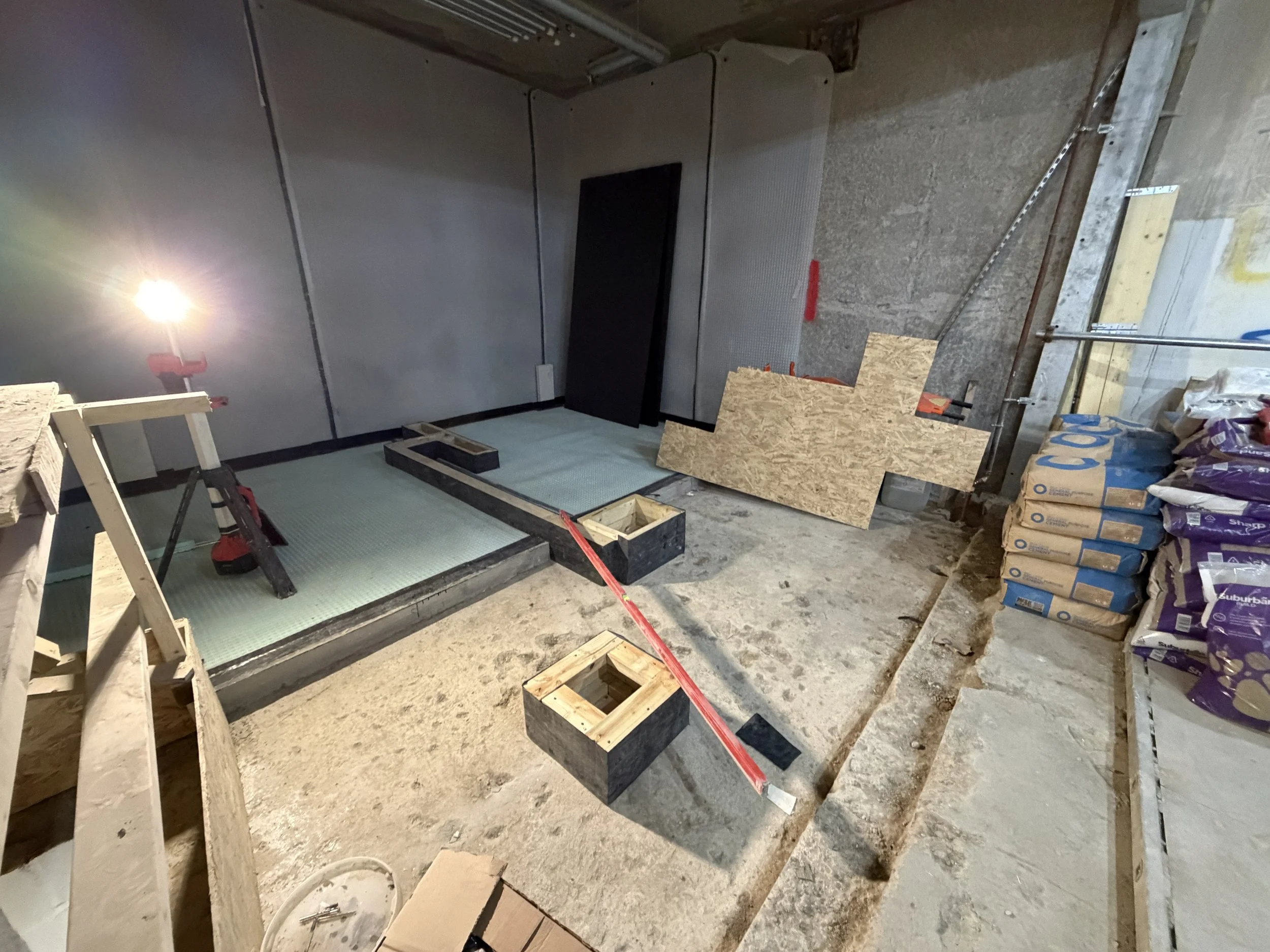Construction site with unfinished flooring, wooden frames, bags of construction materials, and tools including a level and a saw.
