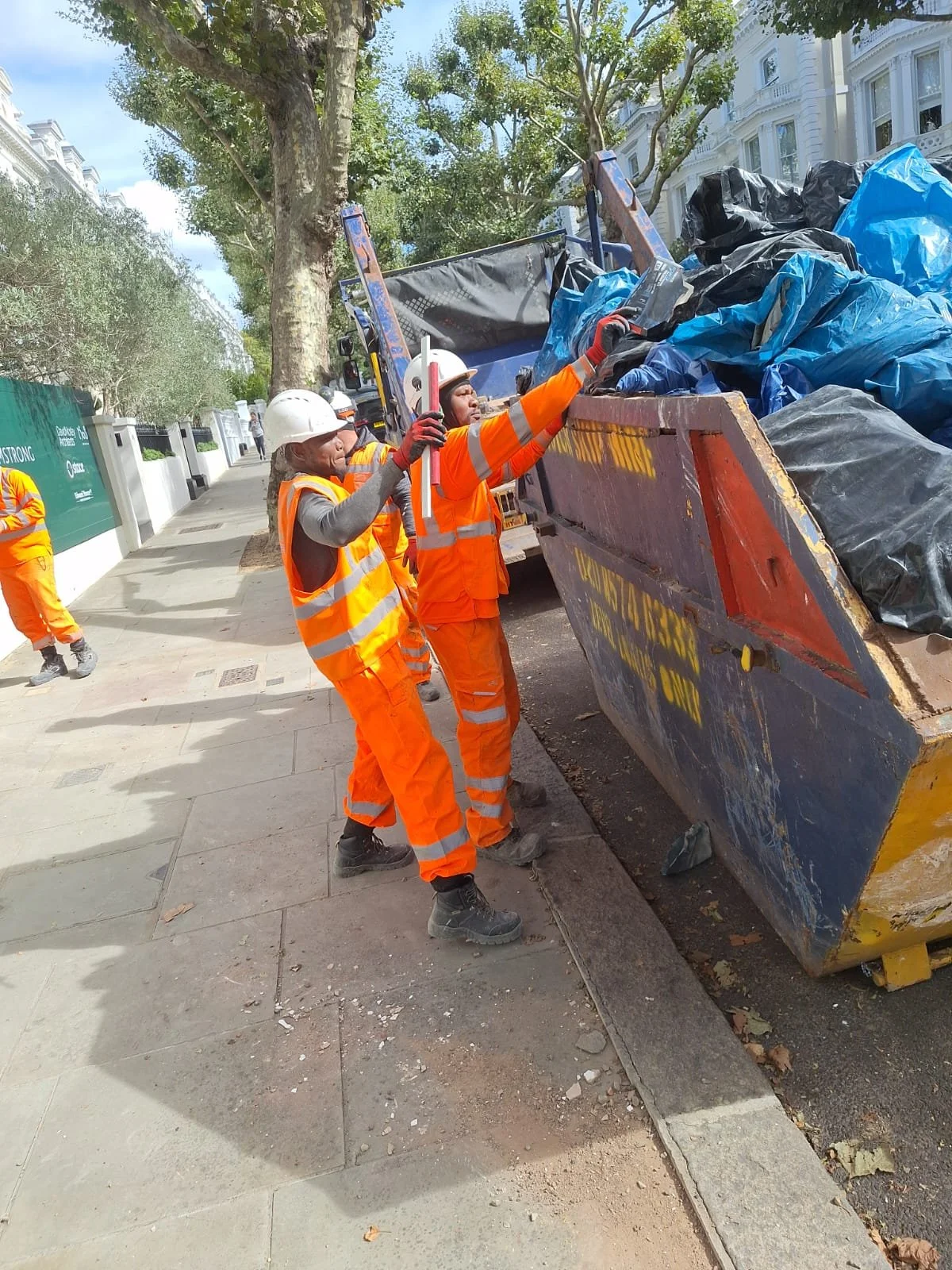 Workers wearing orange safety vests and helmets are unloading trash bags from a commercial trash truck on a city street with trees and buildings in the background.
