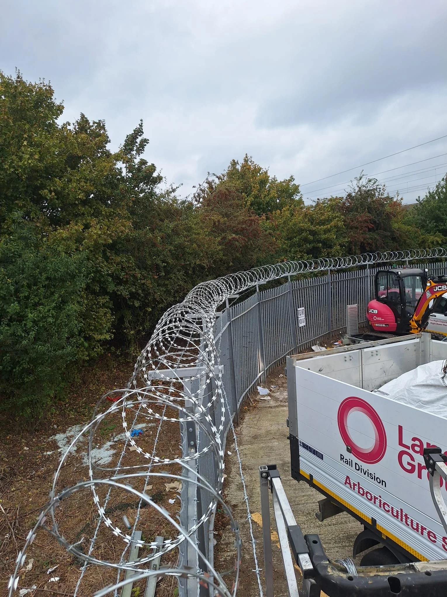 Barbed wire fence alongside construction equipment and a sign, with trees and cloudy sky in the background.