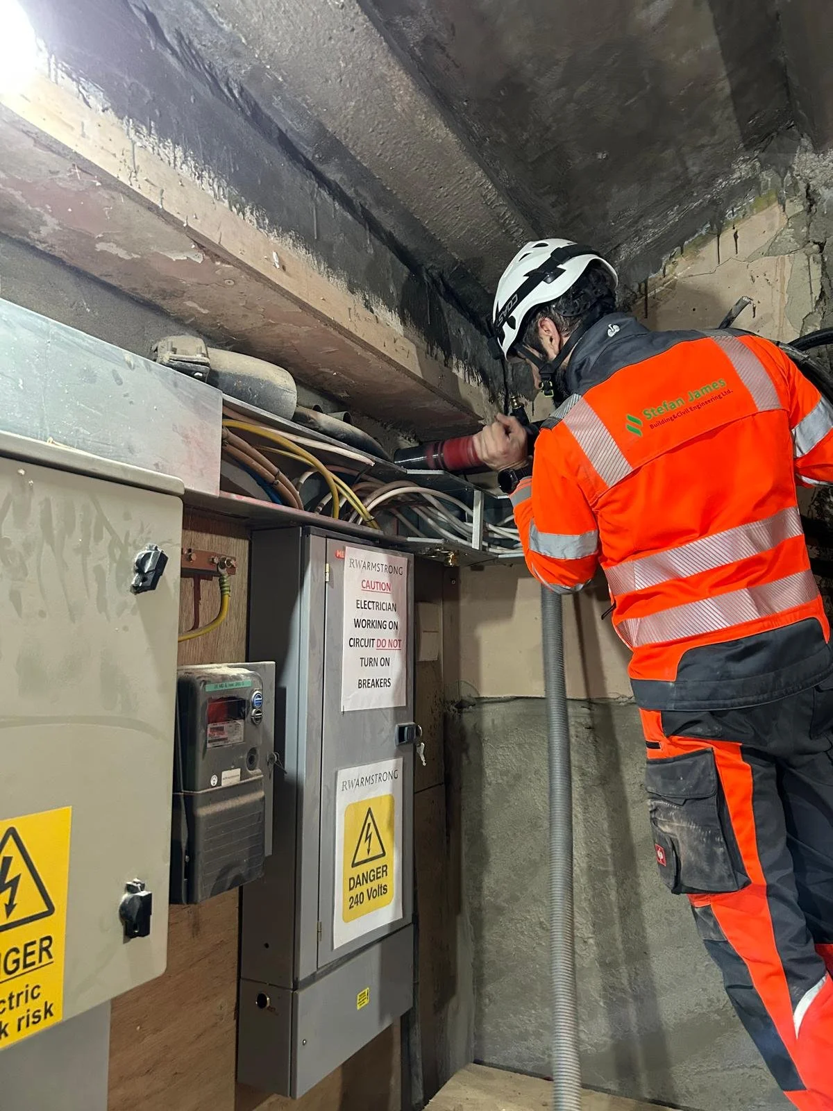 Electrician in safety gear working on electrical wiring in an industrial setting, near electrical panels and warning signs.