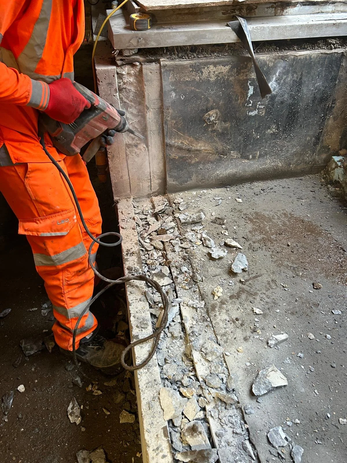 A construction worker wearing orange safety pants, a matching jacket, and red gloves is using a power tool to cut through a concrete wall. There are scattered pieces of broken concrete on the ground, and the workspace appears to be an indoor construction site with visible dirt and debris.