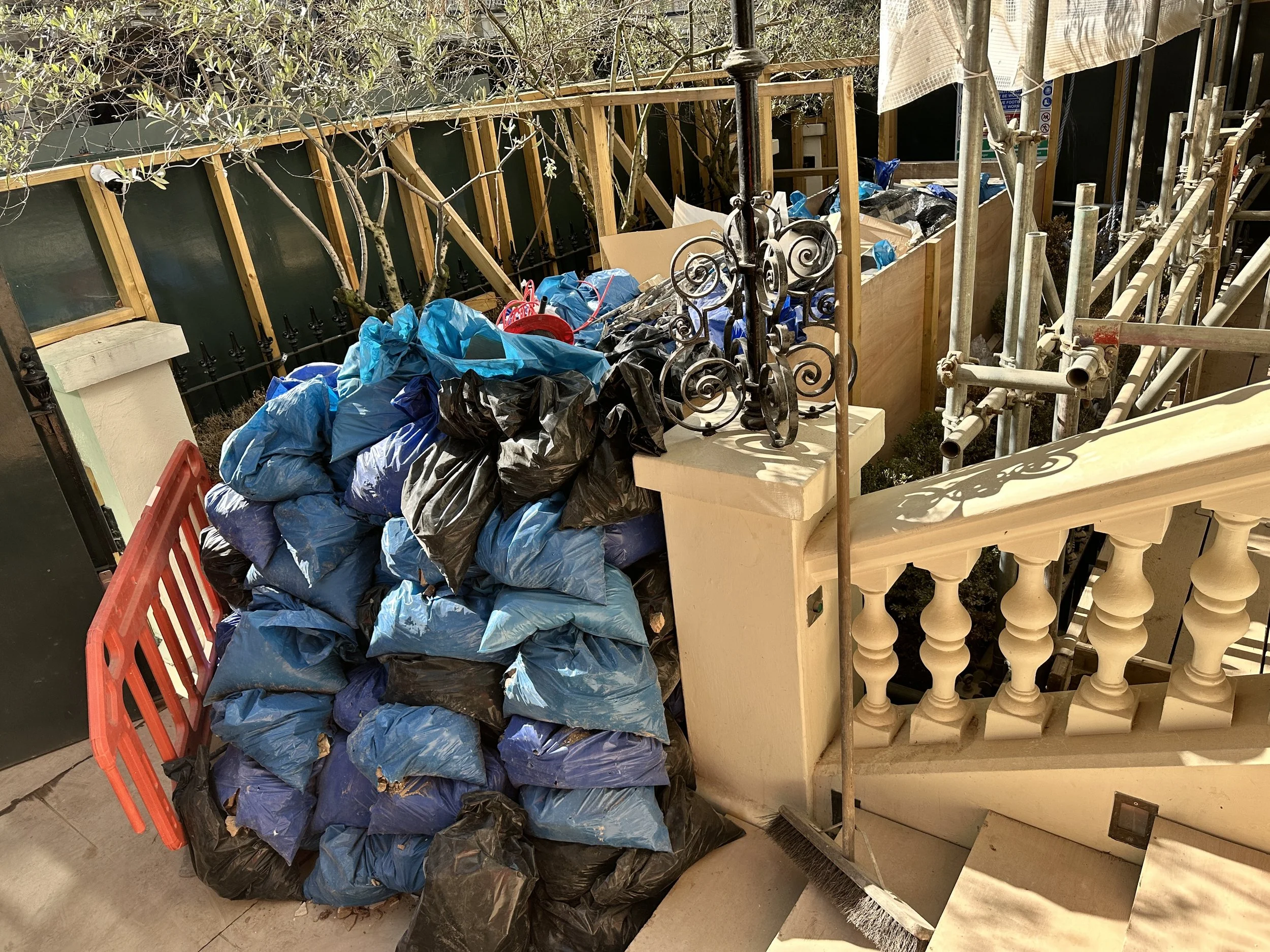 Cluttered area with black and blue trash bags stacked next to a beige decorative balcony railing and a black wrought iron fence with trees and a wooden framework in the background.