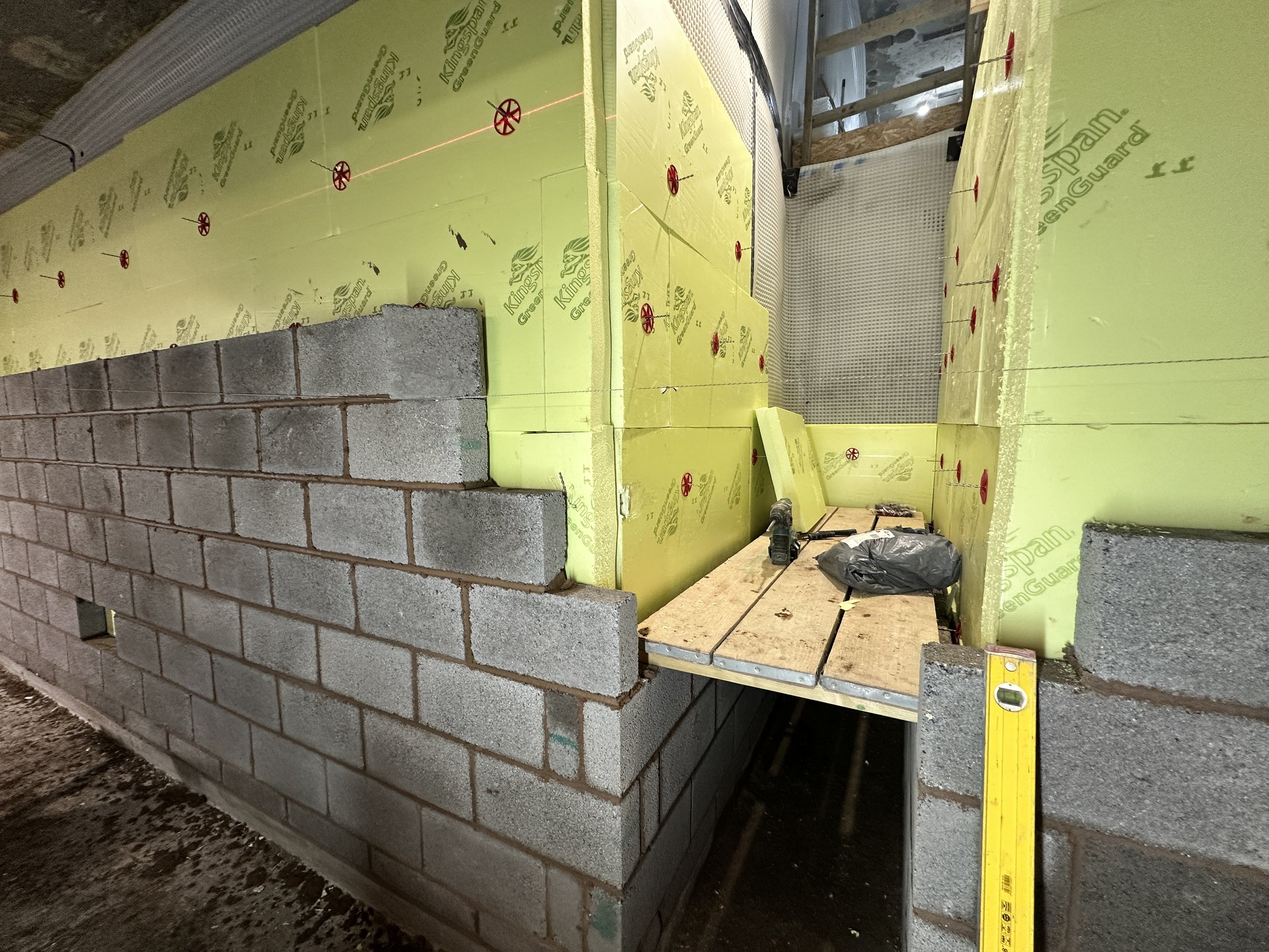 A partially built brick wall with gray cinder blocks on a construction site. Yellow insulation foam boards are placed behind the wall, secured with red plastic fasteners. Construction tools such as a level tool and a drill are visible on the floor near the wall. Wooden beams and other construction materials are also present in the background.