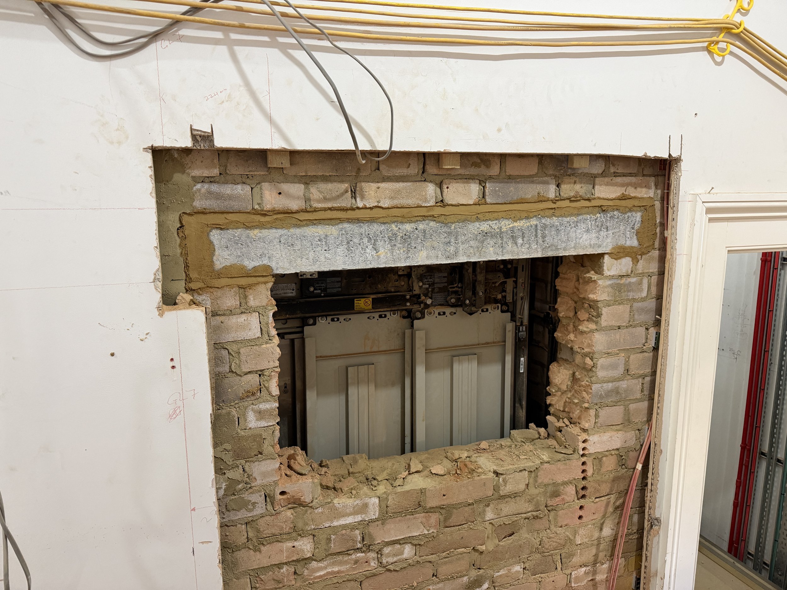 Construction site with an opening in a brick wall revealing the interior of an elevator shaft with exposed wiring and electrical components.