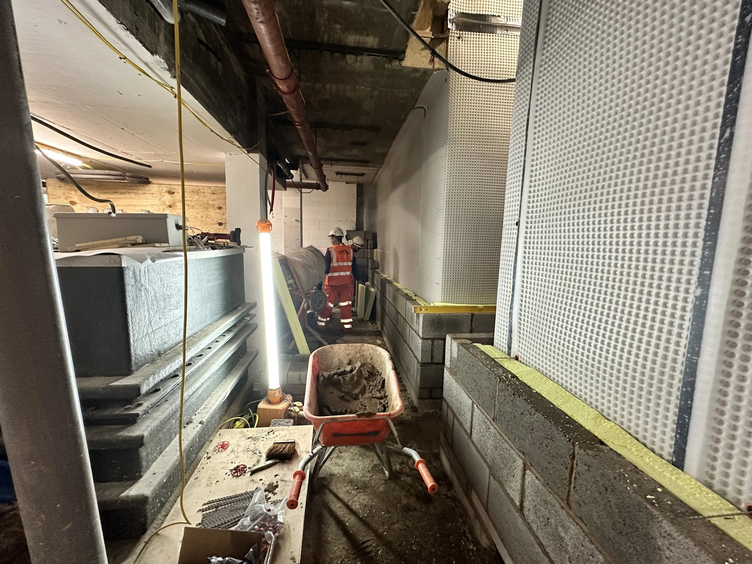Construction site with workers in safety gear installing or working on a wall with perforated panels. Construction tools and materials are scattered around, including a wheelbarrow filled with dirt or cement and a workbench with various items.