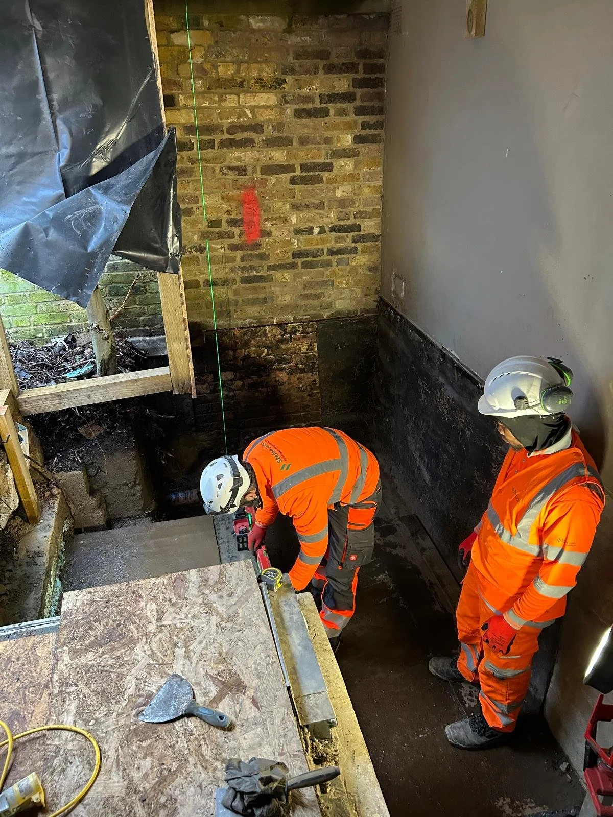 Two construction workers wearing orange safety suits, helmets, and gloves working in a basement or foundation area with bricks and concrete.