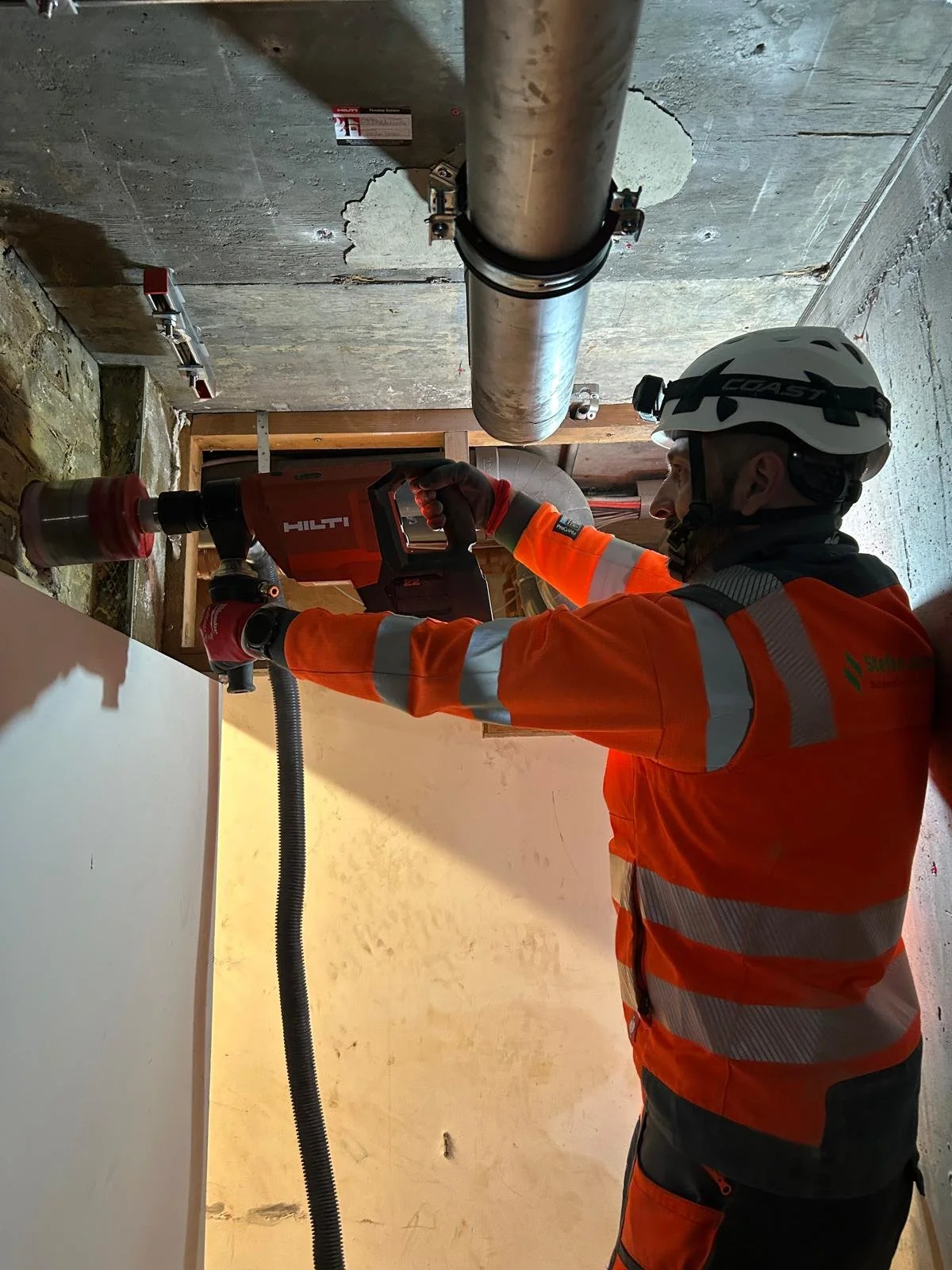 Worker in orange safety gear and helmet using a Hilti power drill to secure a pipe under a concrete ceiling.