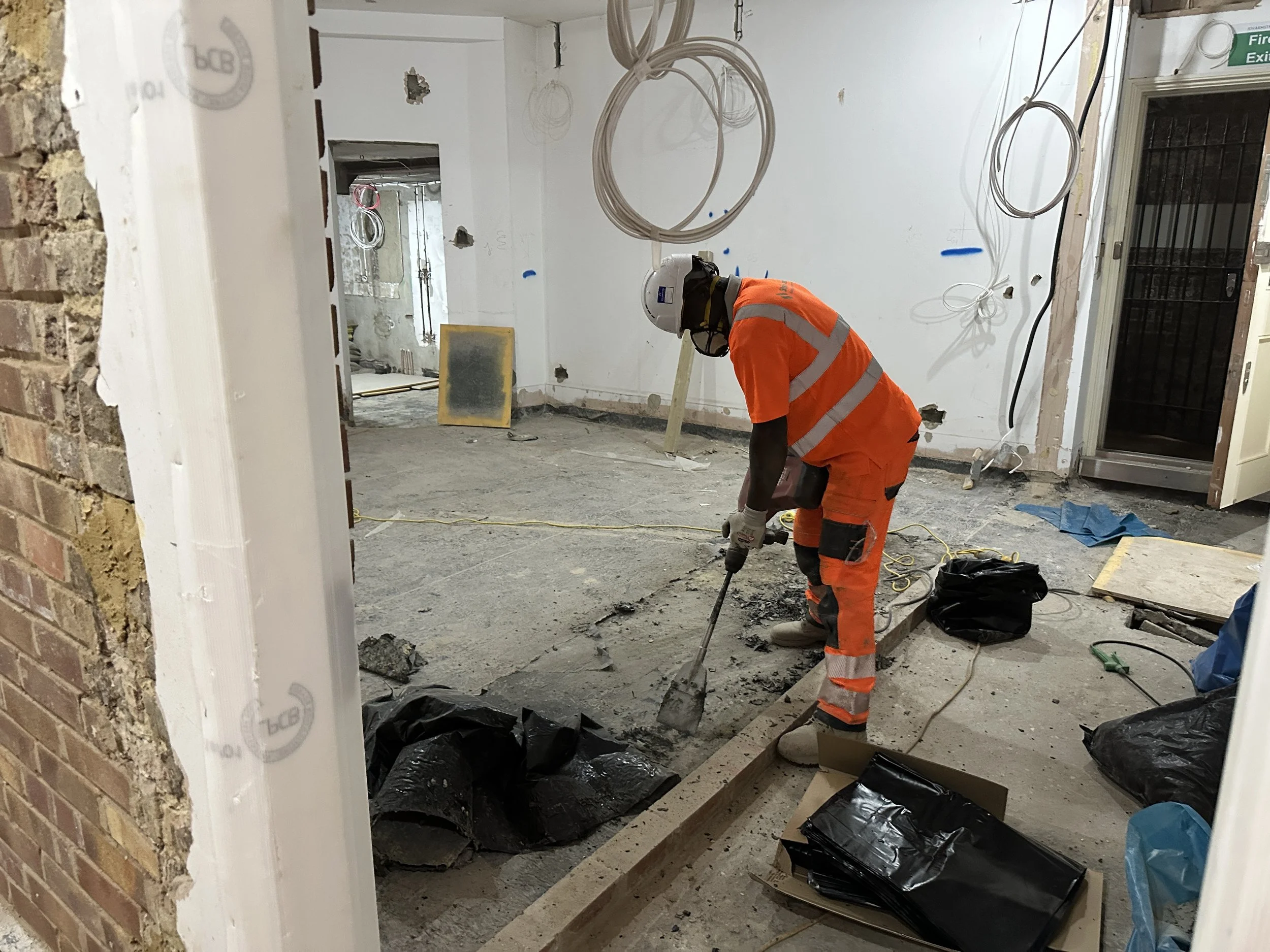Construction worker wearing an orange safety vest and helmet uses a jackhammer to break concrete floor inside a building under renovation. The room has construction debris, wires hanging from the ceiling, and partially finished walls.