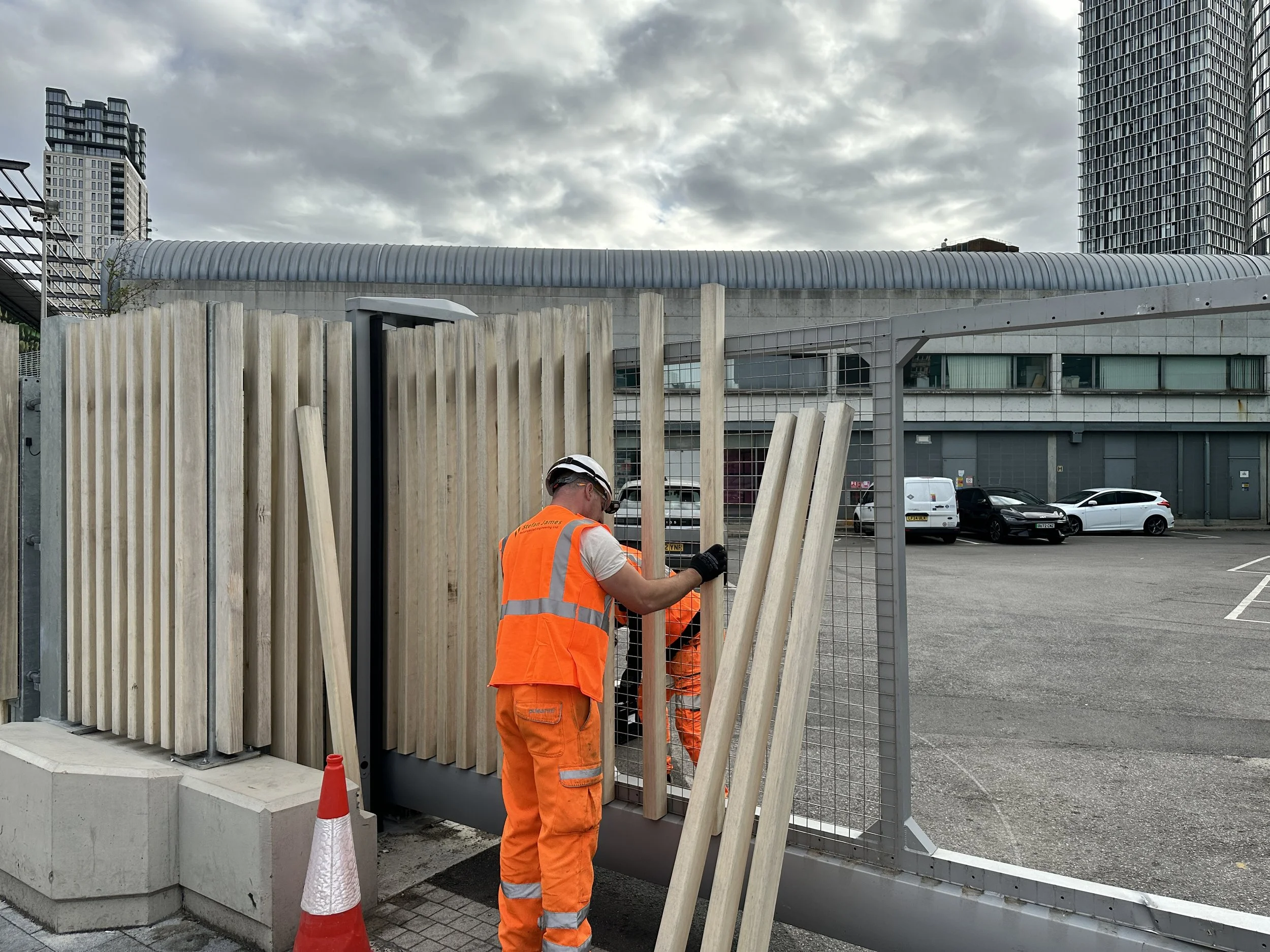 Construction workers installing a wooden fence at a parking lot with tall buildings in the background.