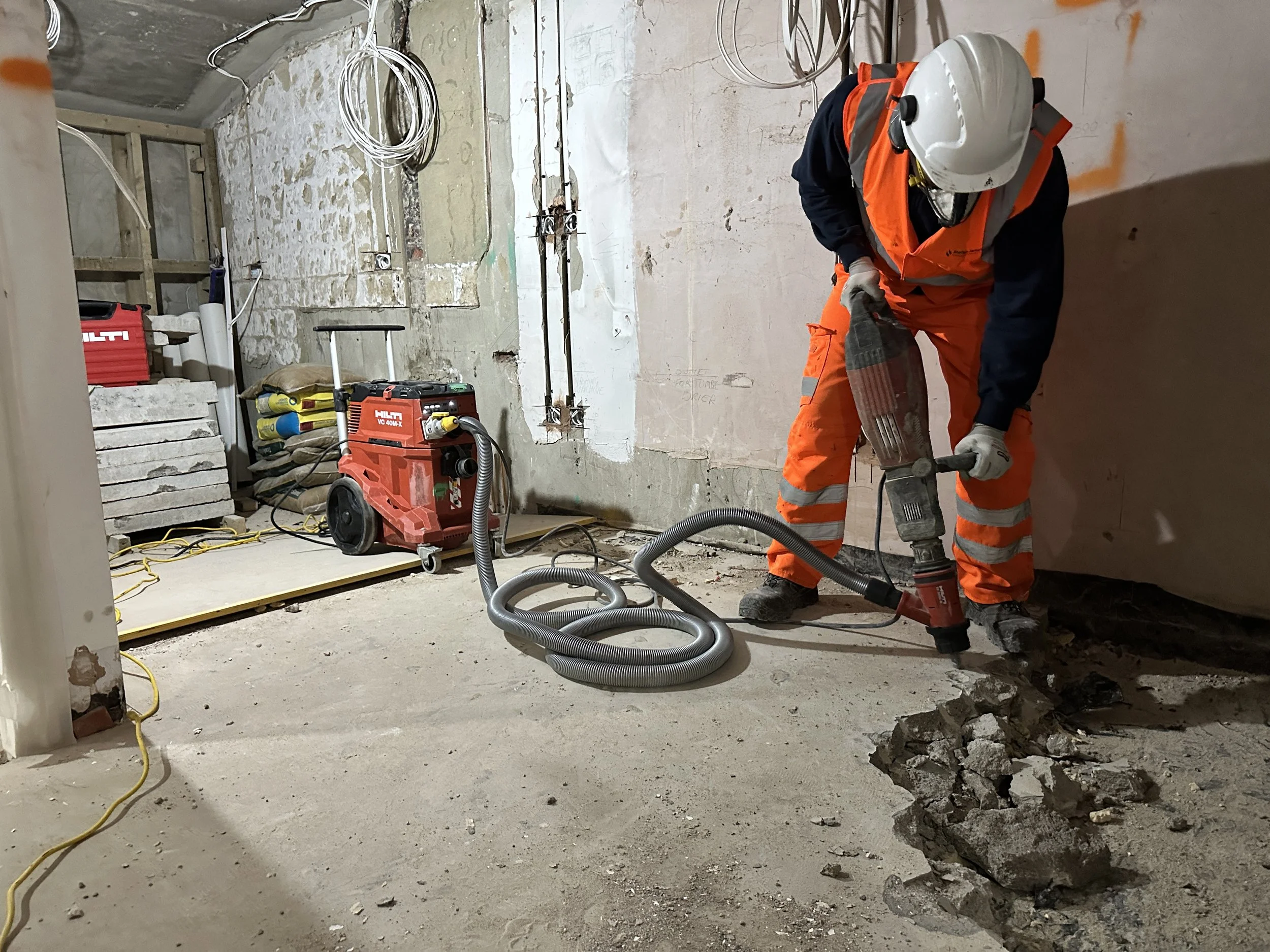 Construction worker wearing orange safety vest, white hard hat, and gloves is kneeling on the floor, working on a construction site with exposed wiring, pipes, and damaged concrete floor.