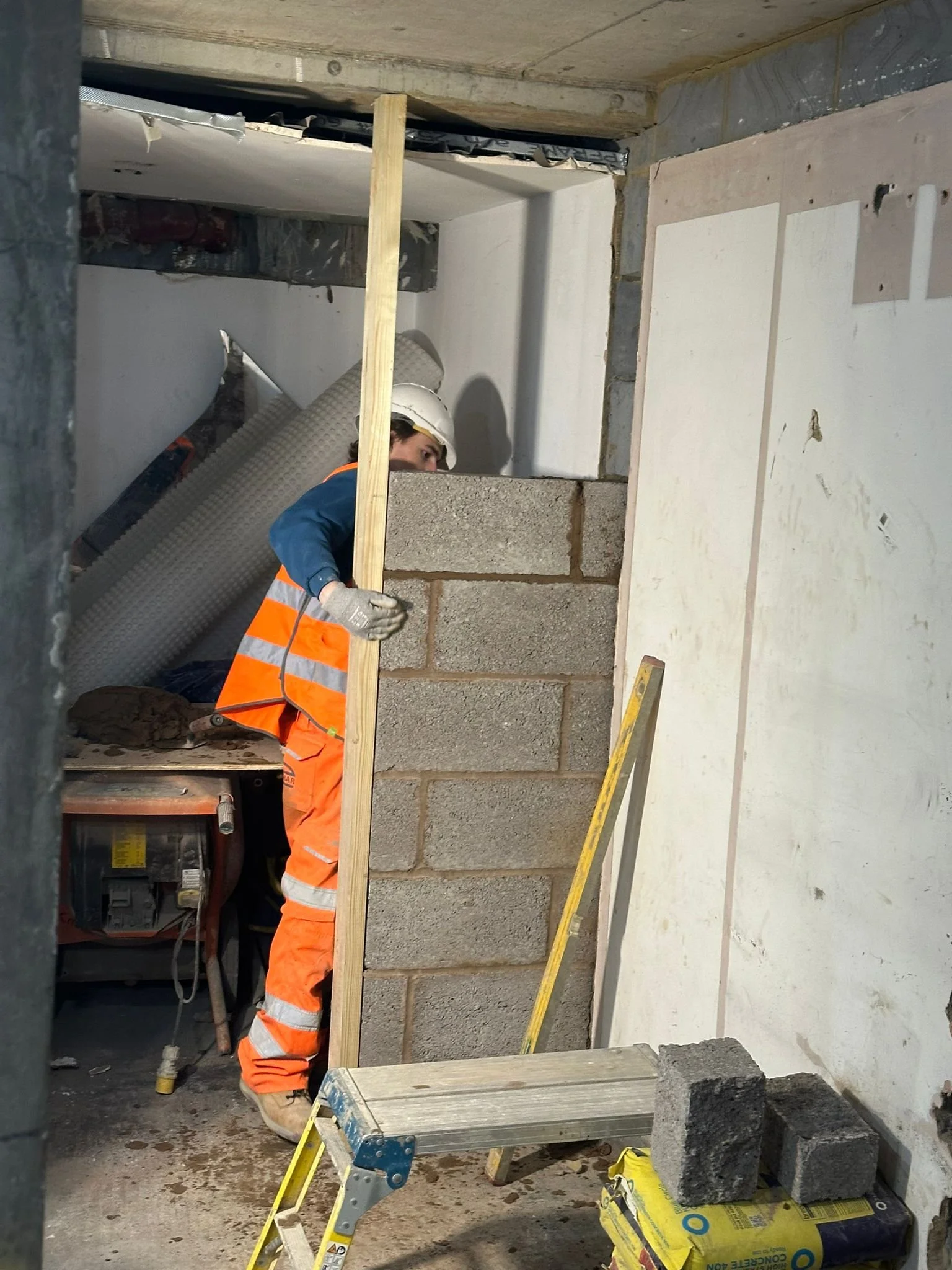 A construction worker wearing an orange safety vest and white helmet is working on a wall, laying bricks with mortar. There are construction materials and tools around, including a yellow level, a stack of cement blocks, and a ladder.