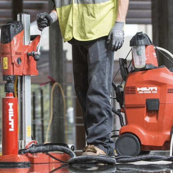 Person operating a red hydraulic tool on a construction site, with a red pressure washer nearby.