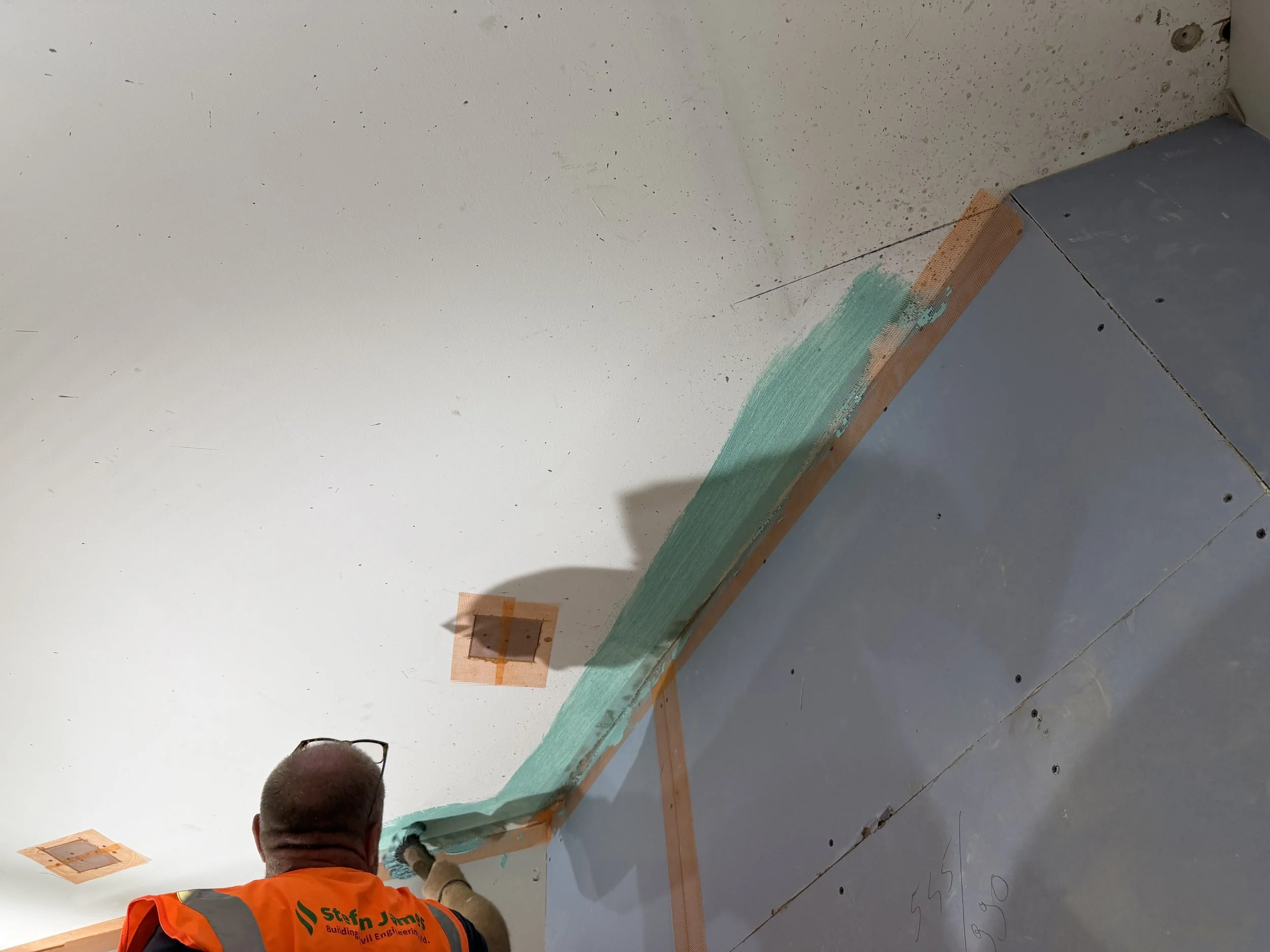 A construction worker with glasses, wearing an orange safety vest with green lettering, working on installing a foam insulation panel on a wall or ceiling, with taped seams and a partially finished surface.