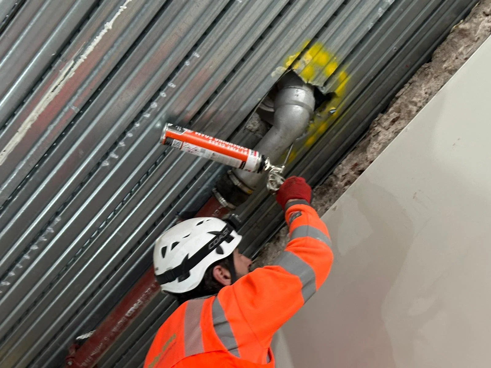 A worker in an orange safety jacket and white helmet uses a caulking gun to apply sealant around pipes on a ceiling with exposed metal ductwork.