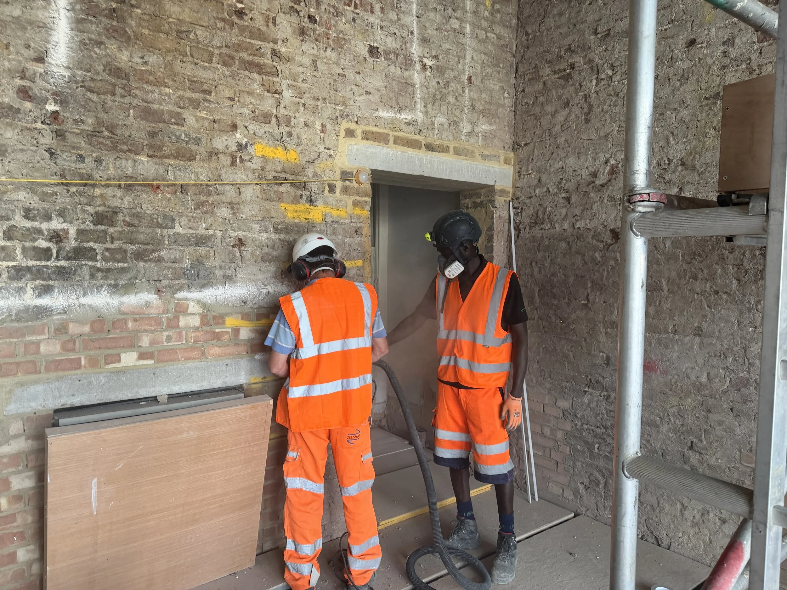 Two construction workers in orange safety vests and hard hats working on a brick wall with scaffolding nearby.