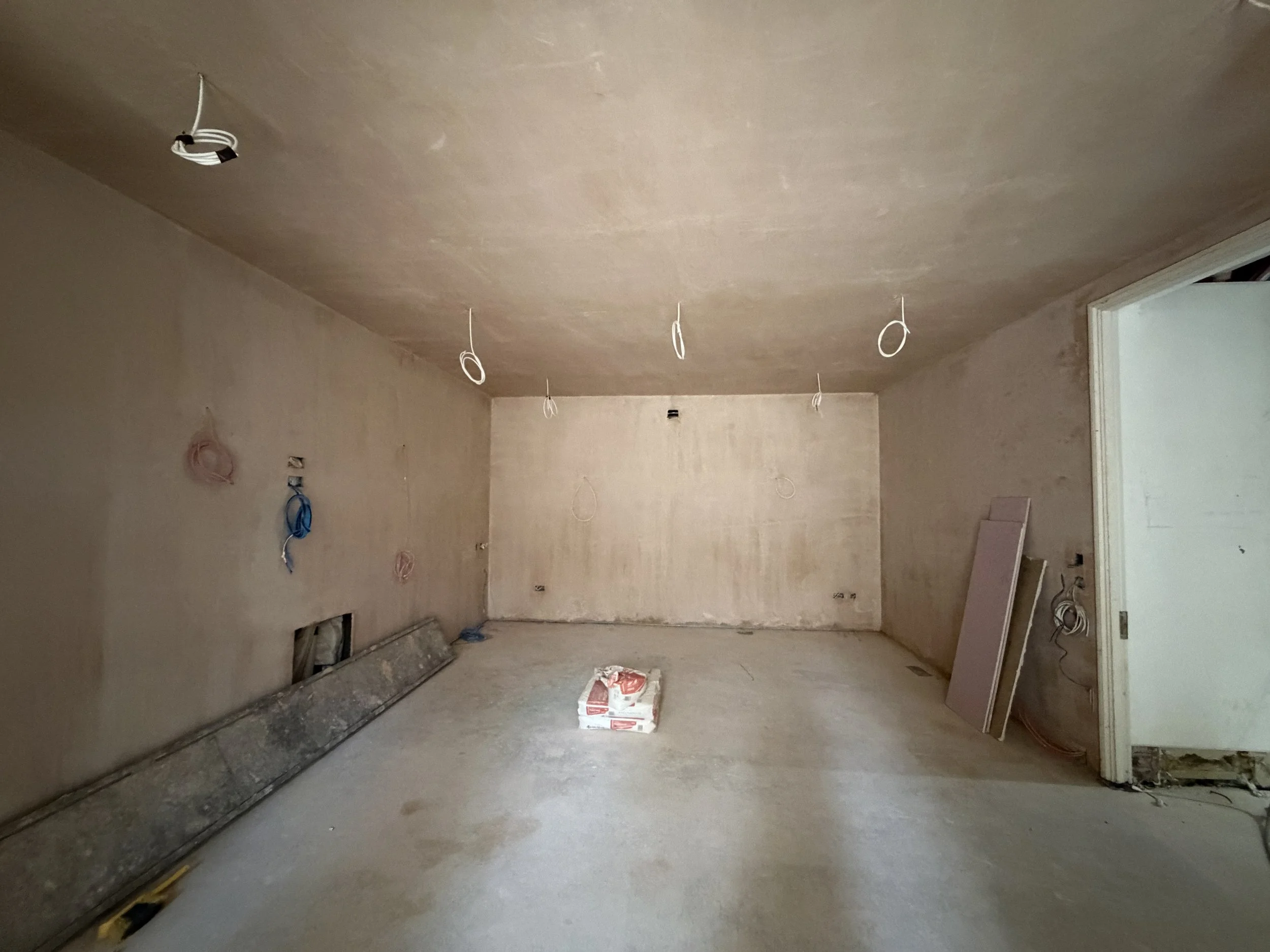 An unfinished room under construction with electrical wiring hanging from the ceiling and walls. There is a large bag of construction materials on the floor, and some drywall panels leaning against the wall.