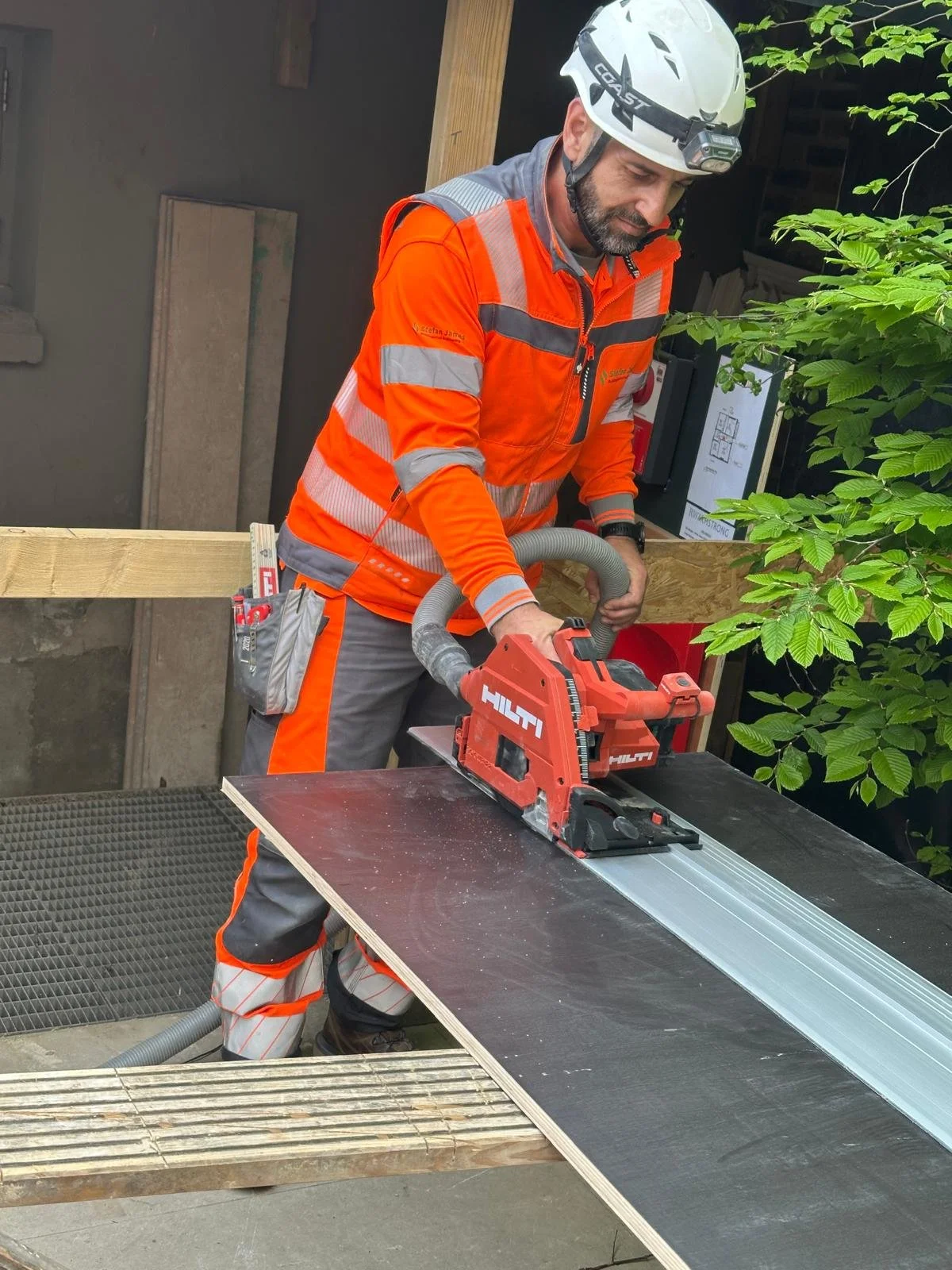 A construction worker wearing a white safety helmet, orange high-visibility jacket, and gray work pants using a red Hilti power saw on a piece of black wood, with wood and building materials around him.