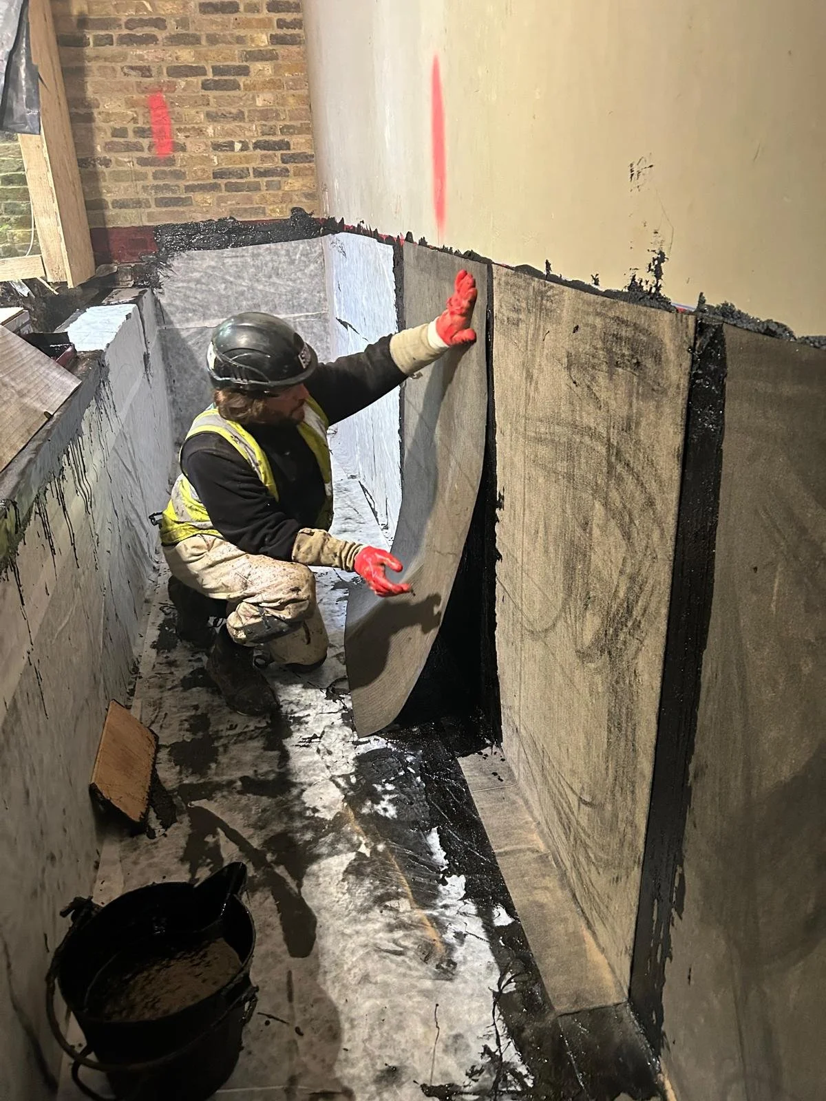A construction worker kneels on the floor, installing a large sheet of waterproof membrane in a trench, with bricks and construction materials around.