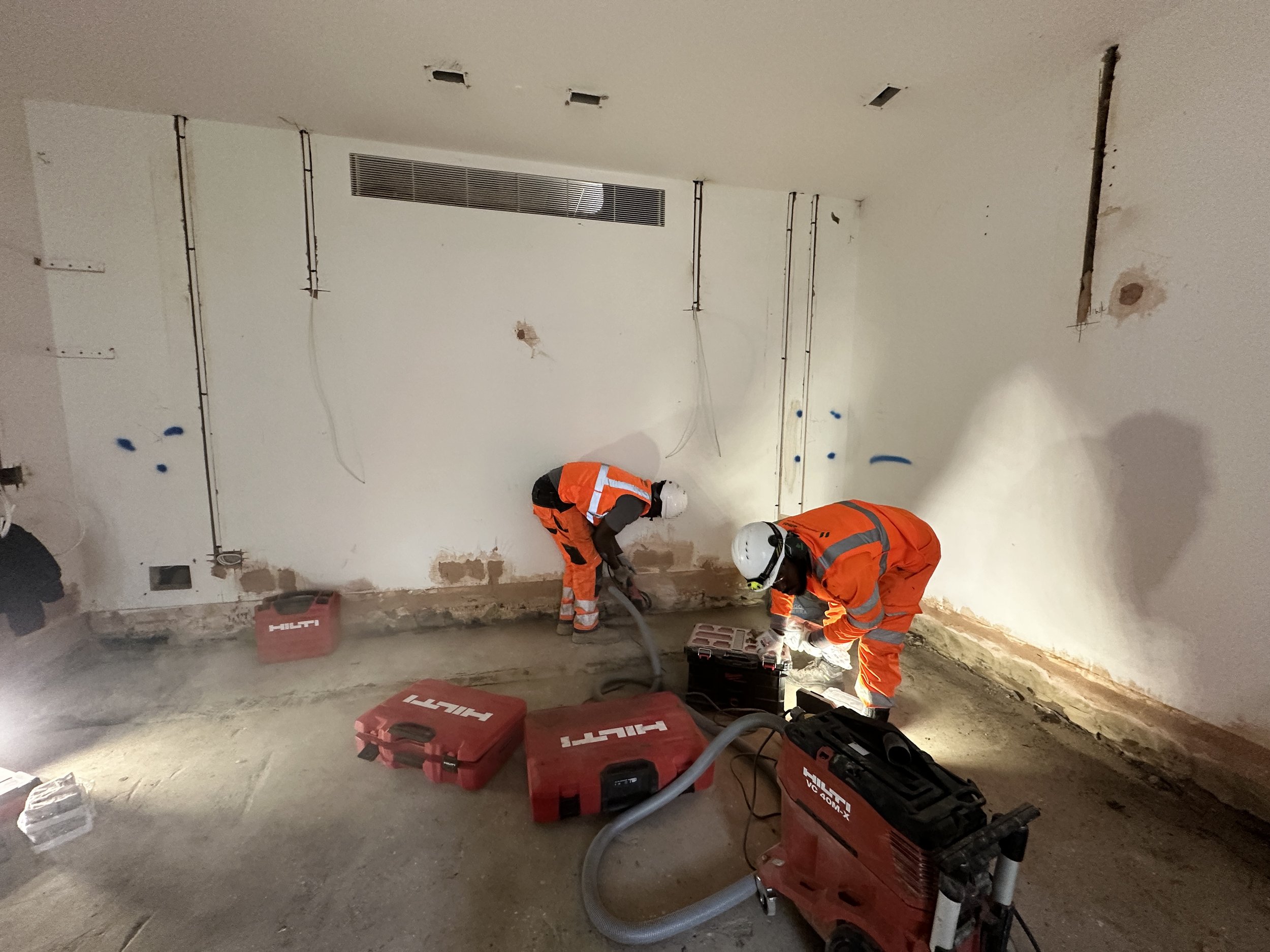 Two construction workers wearing orange safety uniforms and helmets working on wall renovation inside a building. Construction tools and equipment, including red Hilti boxes and a dust extraction vacuum, are visible on the floor.