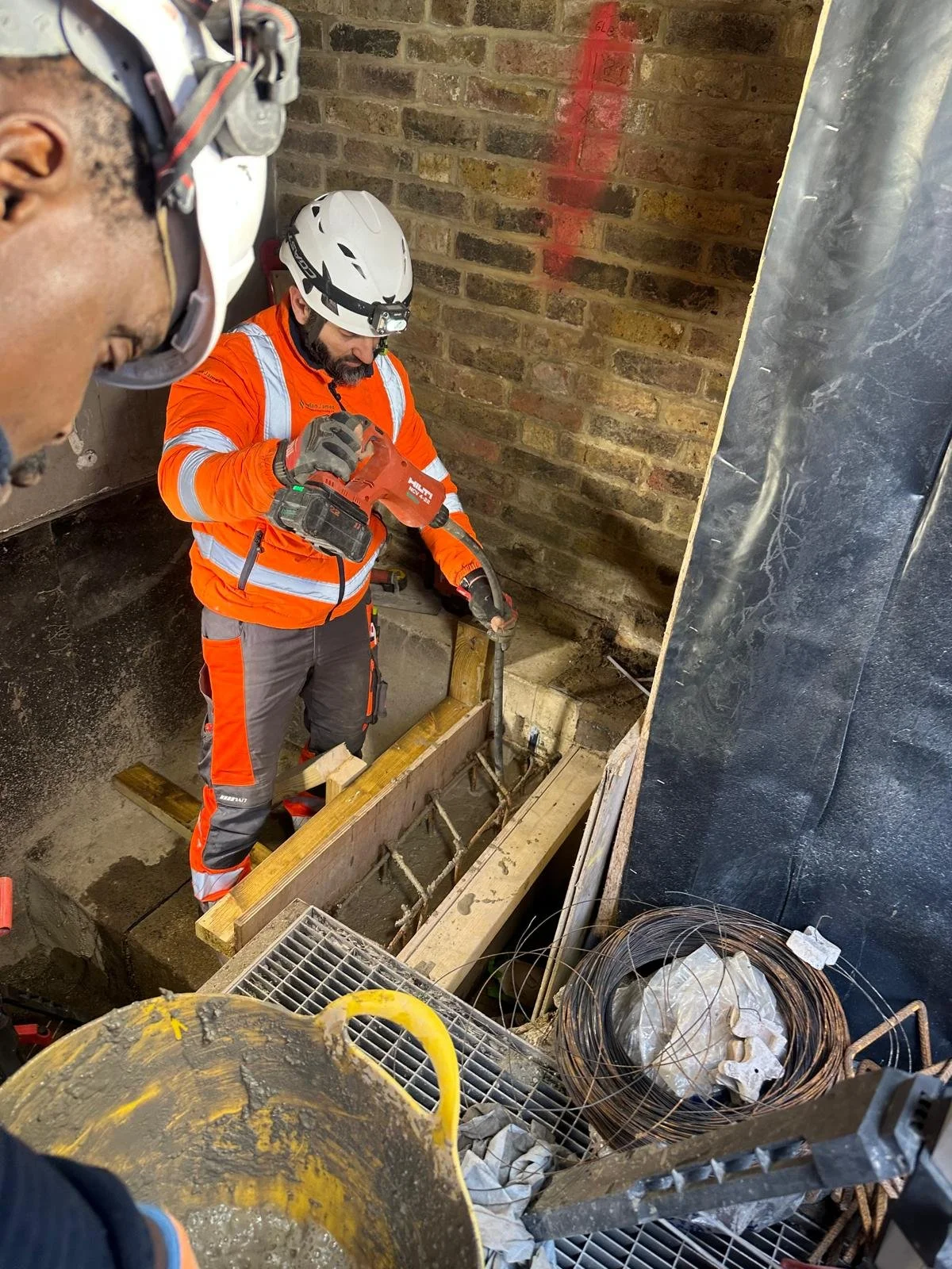 Construction workers performing underground work, one using a power tool, in a small excavation with a brick wall background and construction materials around.