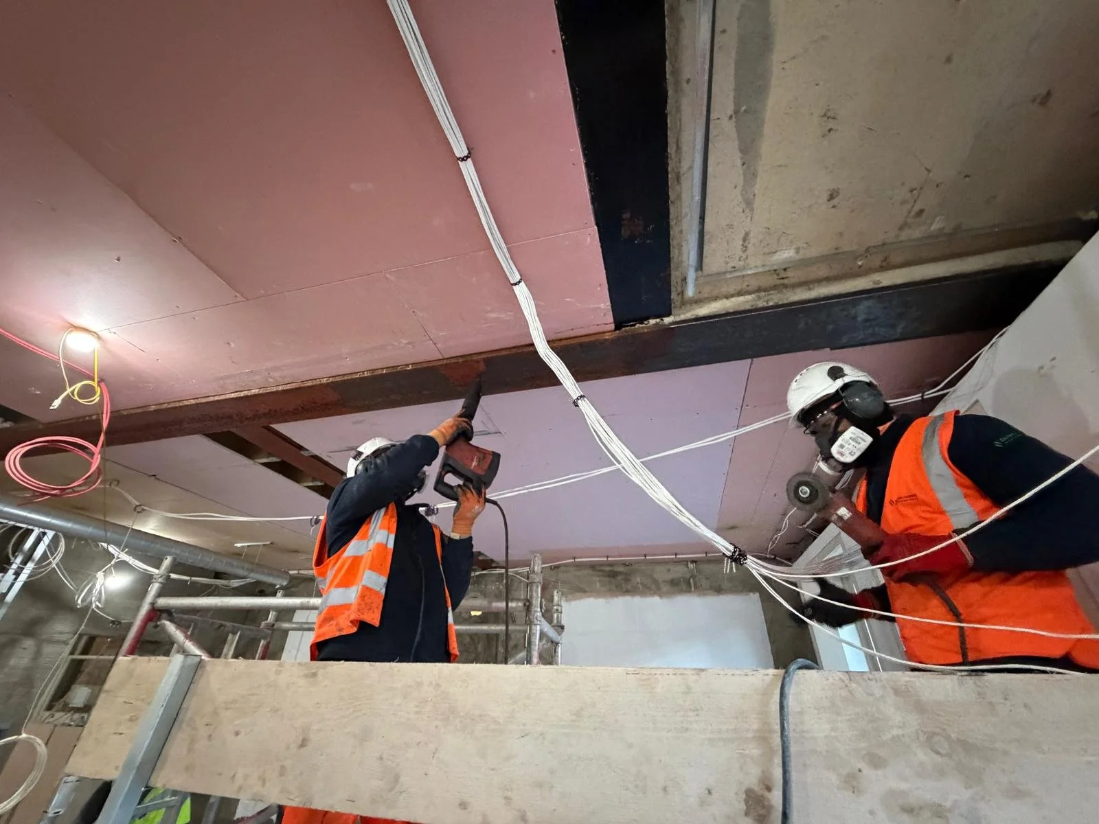 Two construction workers in safety gear and orange vests installing or repairing electrical wiring on a ceiling in a building under construction.