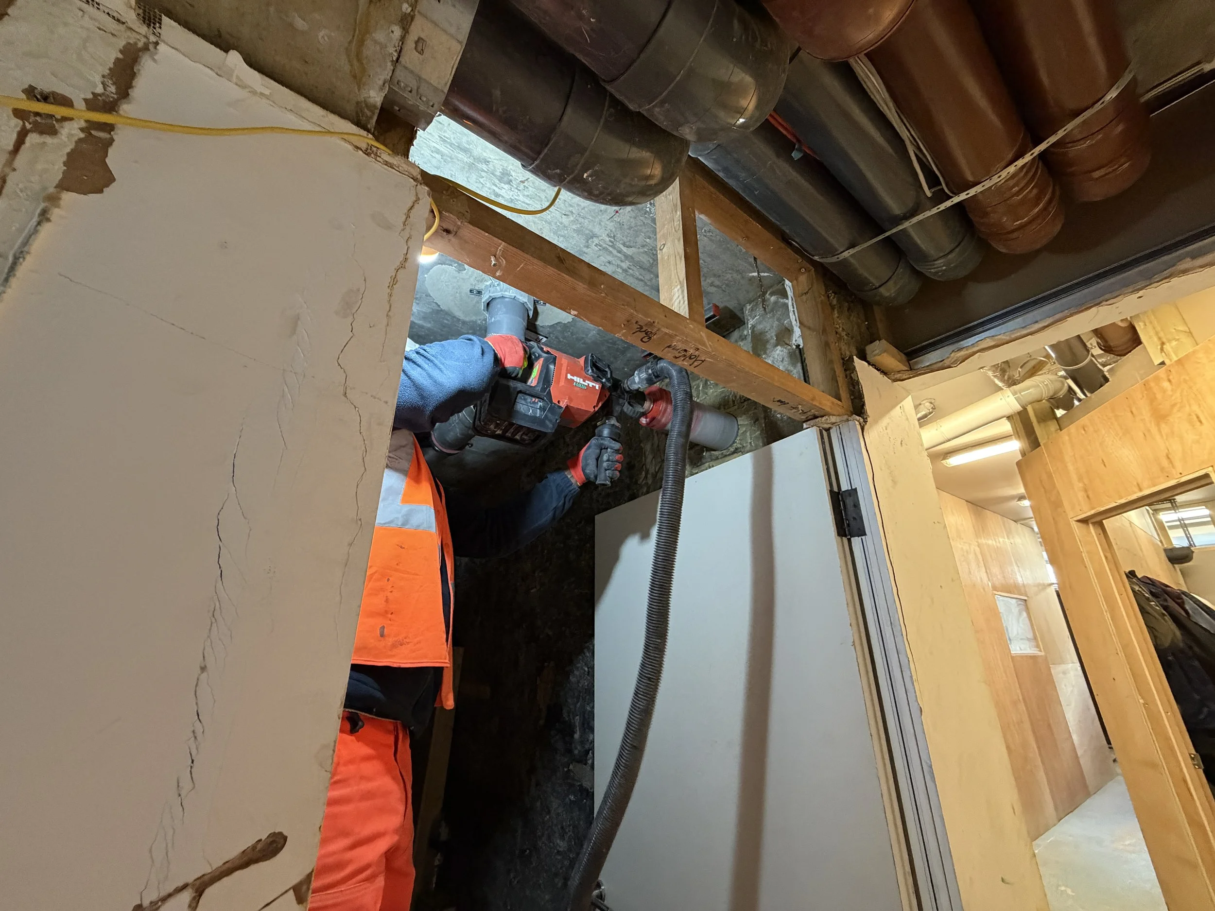 Construction worker using power tools on a construction site with exposed ductwork and pipes overhead.