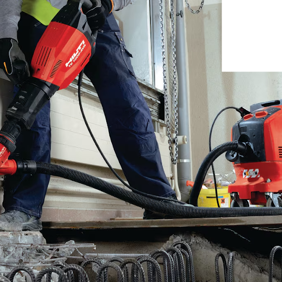 Person using a red and black industrial vacuum or blower to remove debris from a construction or repair site, with tools and chains visible in the background.