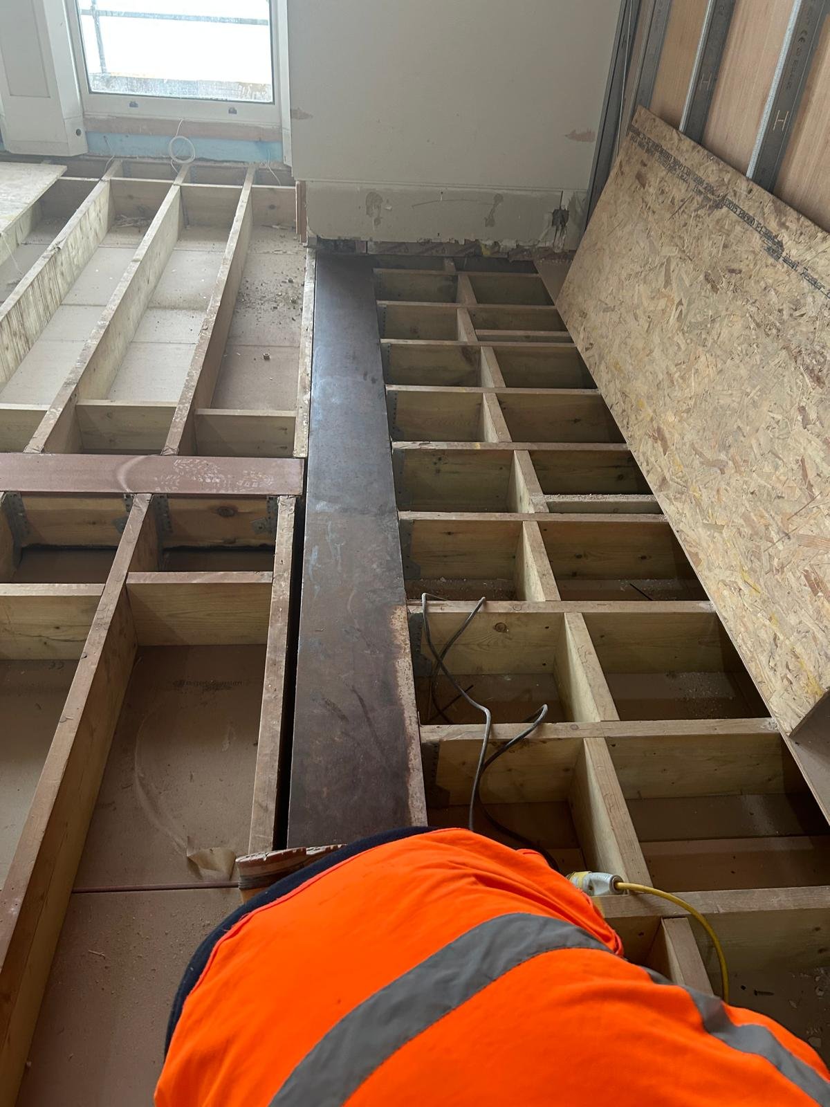 Construction site showing wooden floor framing with some sections covered with OSB plywood, electrical wiring, and a worker in an orange safety vest.