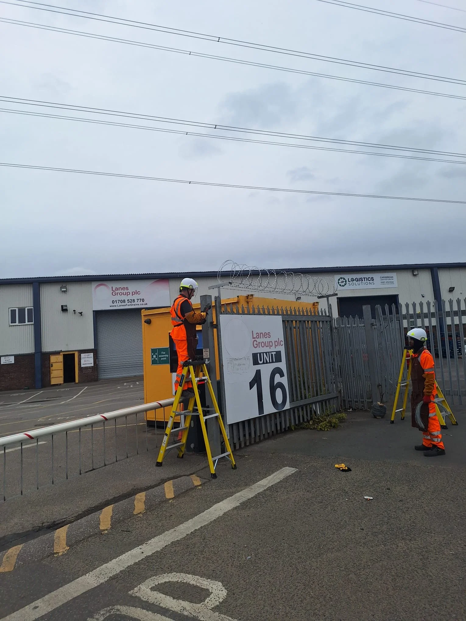 Workers installing or maintaining equipment near a barbed wire fence outside a commercial building, with signage including 'Lanes Group plc' and various industrial elements.