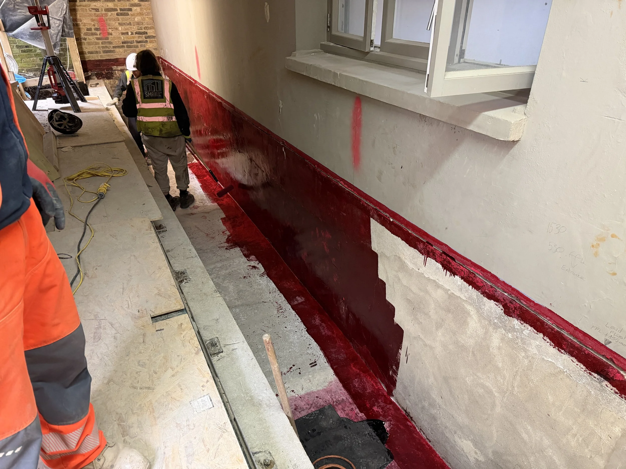 Construction workers painting a wall with dark red paint inside a building. One worker is rolling paint on the lower part of the wall, while another stands nearby. Construction tools, a helmet, and yellow extension cords are visible.