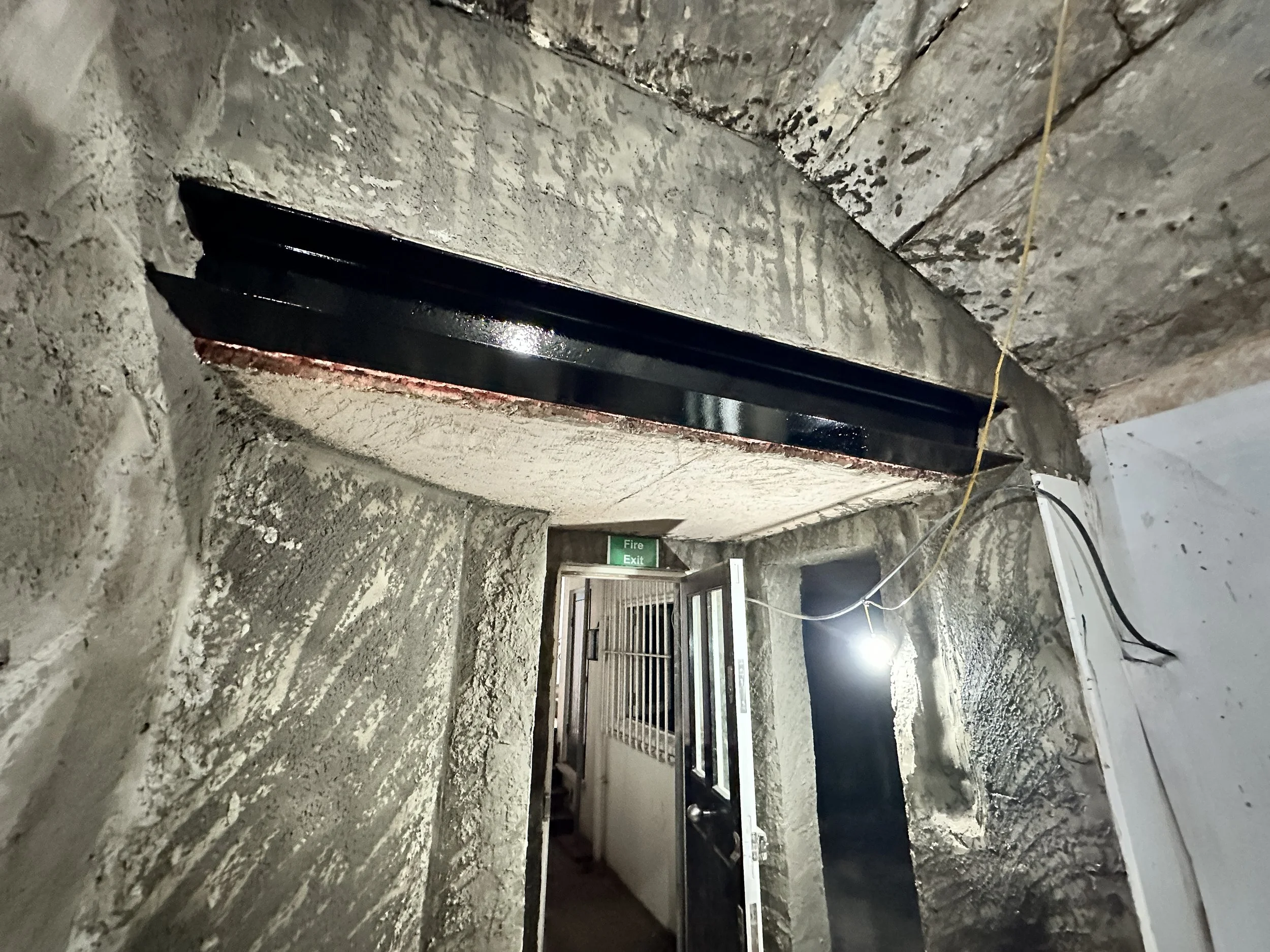 Construction site hallway with rough concrete walls, ceiling, and floor, featuring exposed wiring, a fire exit sign, and scaffolding or metal seating in the background.
