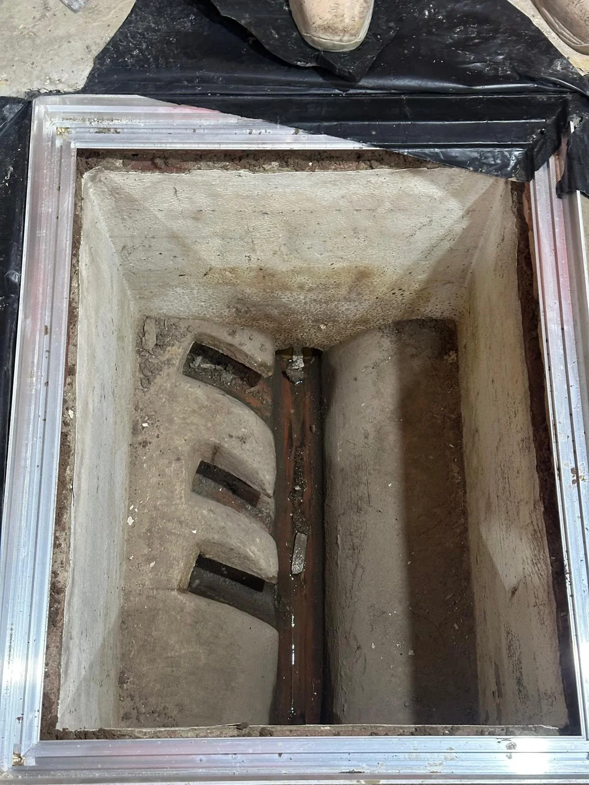 Open concrete trench with embedded drainage pipe and drainage slots, surrounded by construction framing and black plastic sheeting.