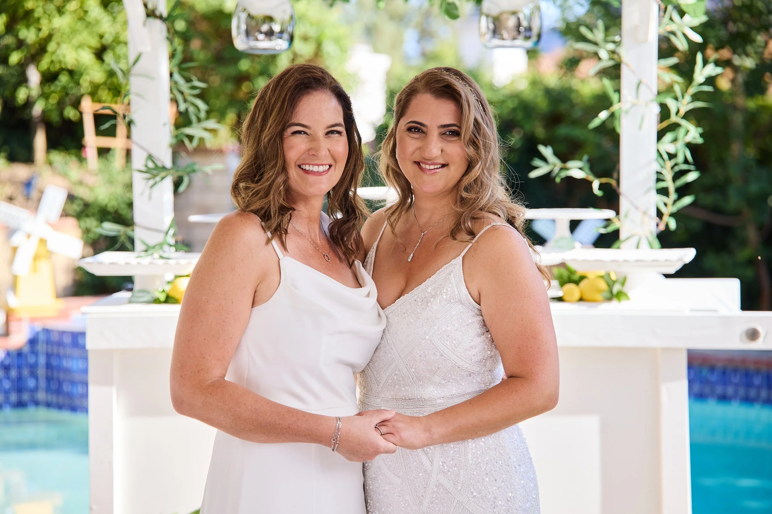 Two women in white dresses holding hands and smiling outdoors during daytime, with greenery and a decorative setup in the background.
