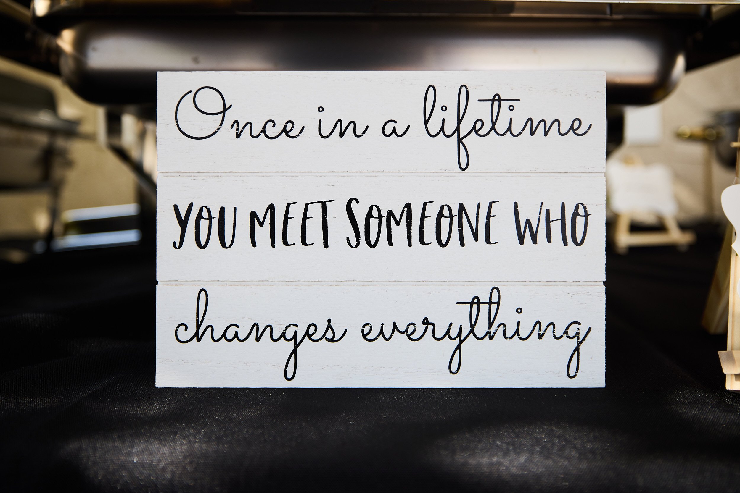 White wooden sign with black cursive and block lettering that reads, 'Once in a lifetime you meet someone who changes everything.'