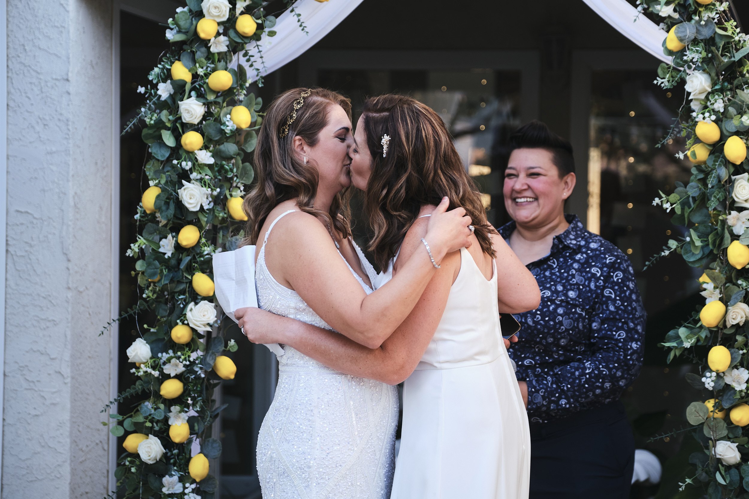 Two women are kissing during a wedding ceremony, with a third woman smiling in the background. They are standing under a floral arch decorated with white roses, yellow lemons, and greenery.