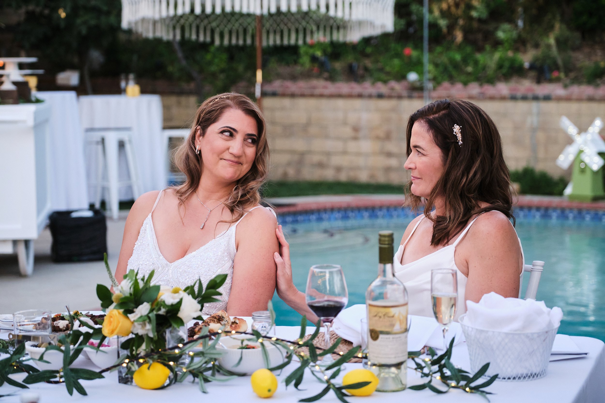 Two women in white dresses sitting at a dinner table by a pool, with one touching the other's arm while they look at each other, during an outdoor celebration.